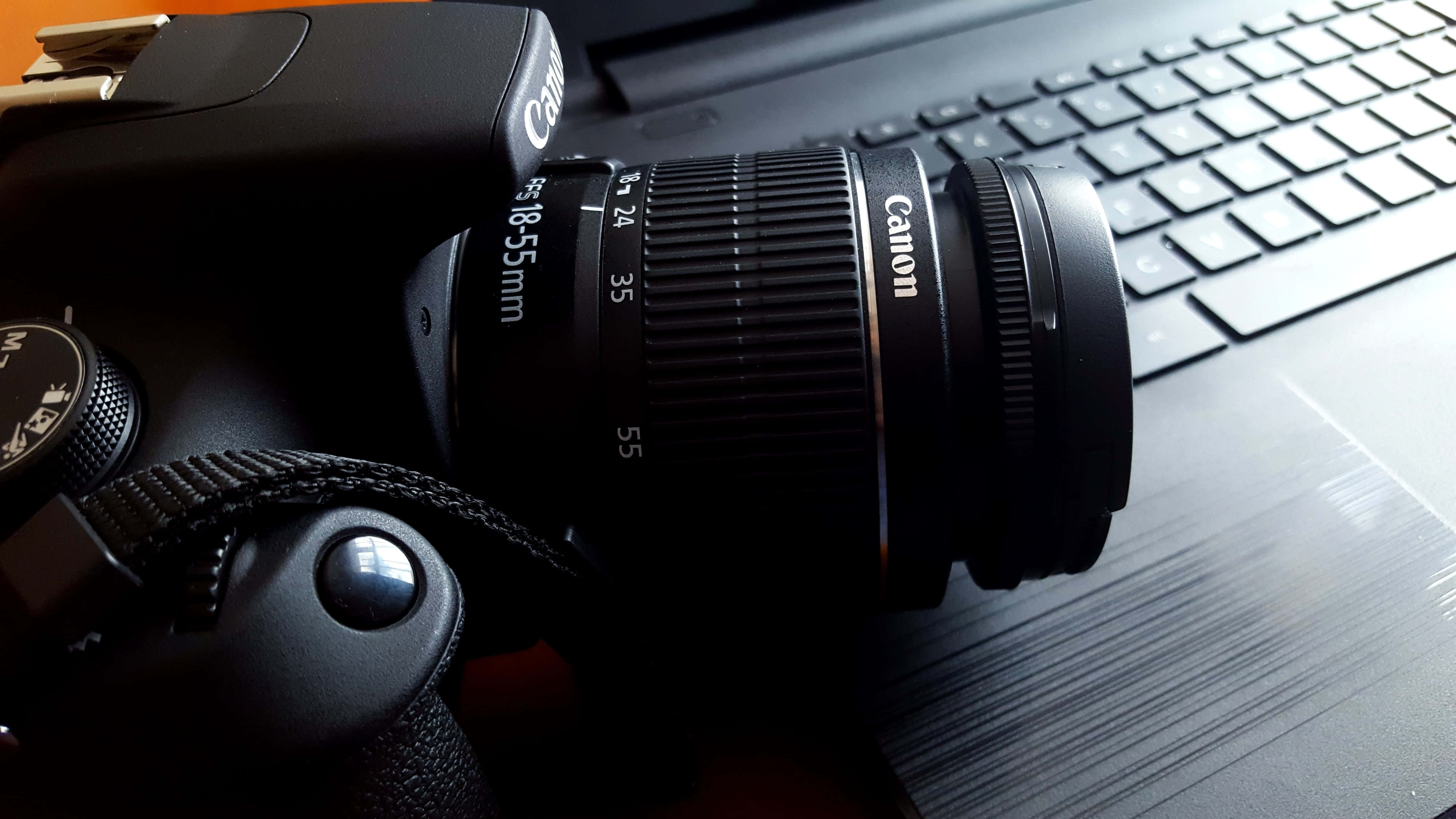 相機 桌布 壁紙 — 一部黑色Canon DSLR相機配備18-55mm鏡頭，放置在昏暗環境中的筆記型電腦鍵盤上。