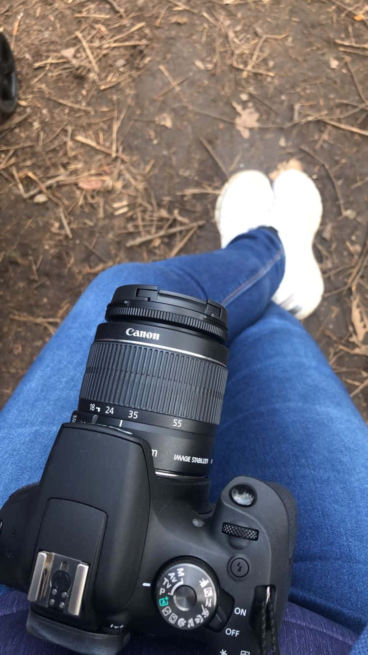 Holding Camera 桌布 壁紙 — 一部黑色Canon DSLR相機放在穿著藍色牛仔褲和白色運動鞋的人的膝上，背景是佈滿乾樹葉和樹枝的森林地面。