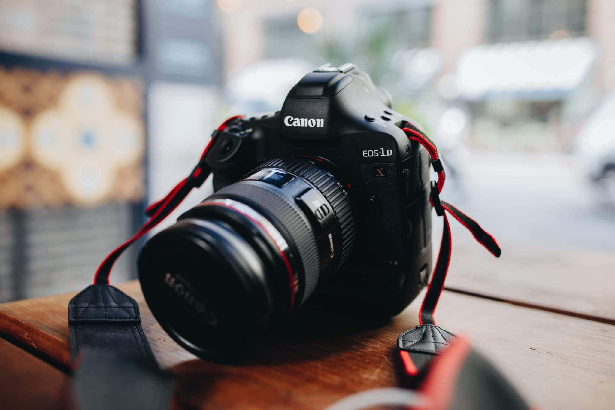 相機 桌布 壁紙 — 一部黑色Canon EOS 1D X DSLR相機搭配紅色相機帶，放置在木桌上，背景是模糊的城市景觀。