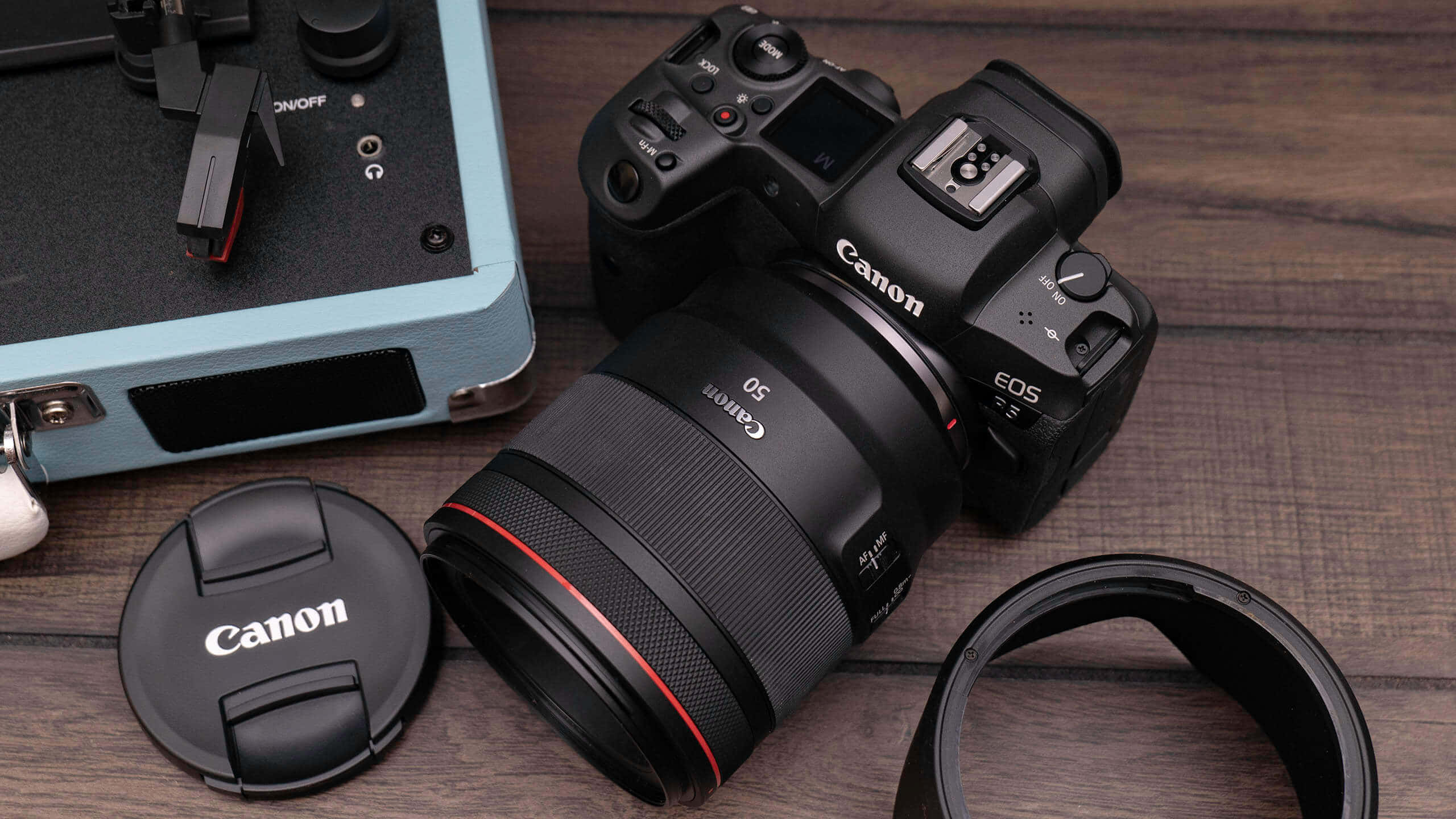相機 桌布 壁紙 — 一部黑色Canon EOS R5相機配50mm鏡頭放在木質表面上，旁邊有鏡頭蓋和淺藍色相機袋。