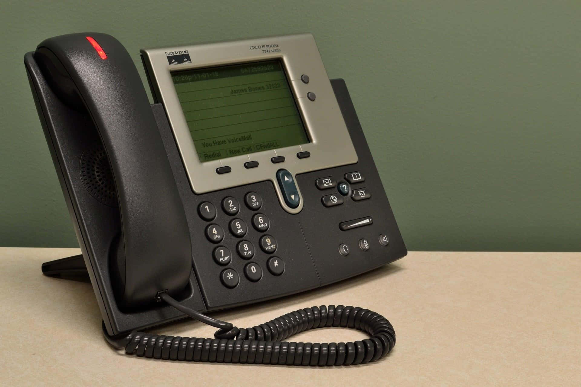 Telemarketing 桌布 壁紙 — 一部黑色Cisco辦公室電話連螺旋電線放在米色桌上，背景是柔和的綠色牆。