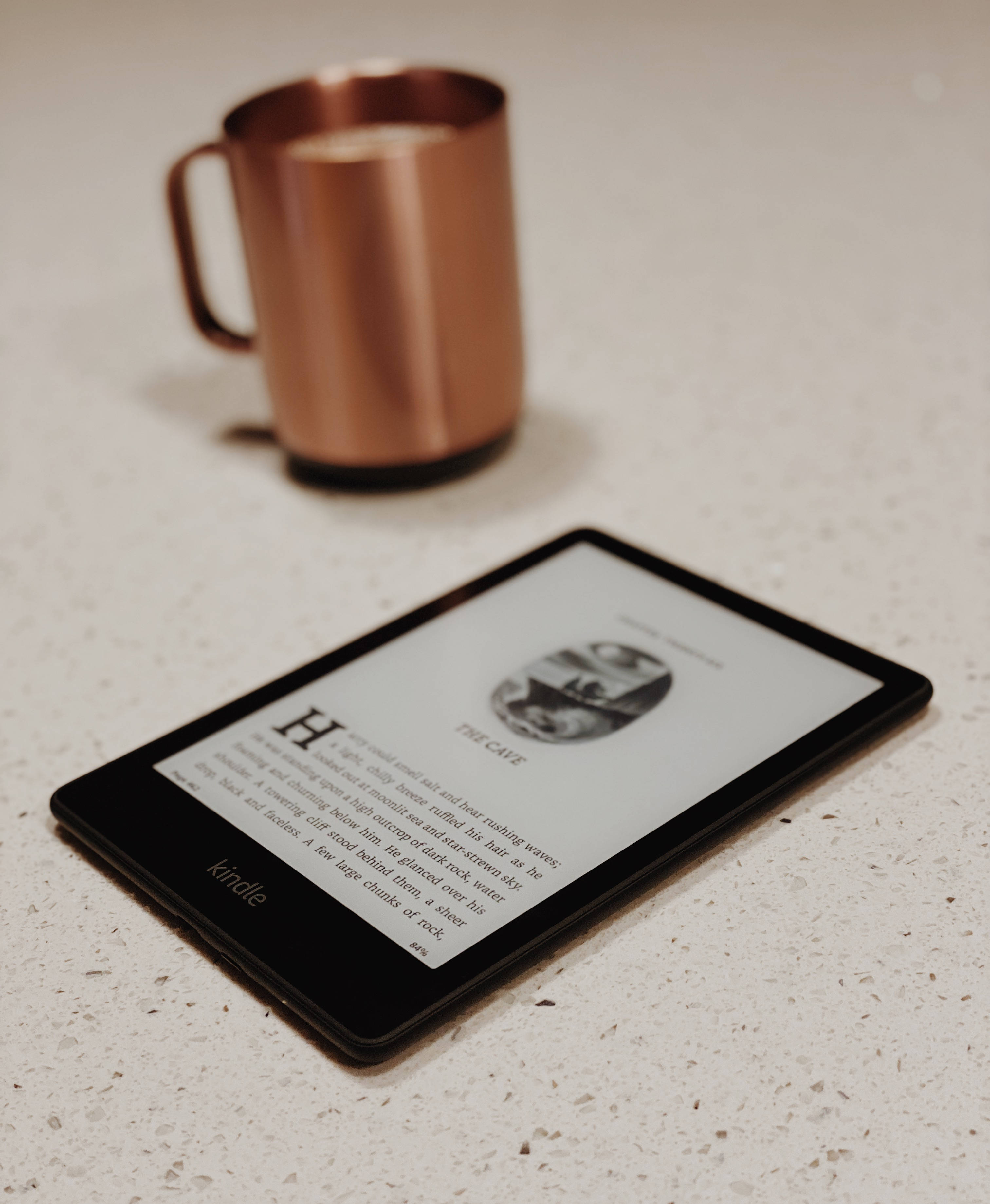dumbledore 桌布 壁紙 — 一部黑色Kindle顯示書頁，放在有斑點的檯面上，旁邊有一個銅杯，在柔和溫暖的光線中。