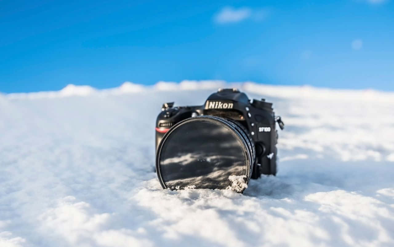 攝影 桌布 壁紙 — 一部黑色Nikon DSLR相機部分埋在純淨的雪中，上方是清澈的藍天。