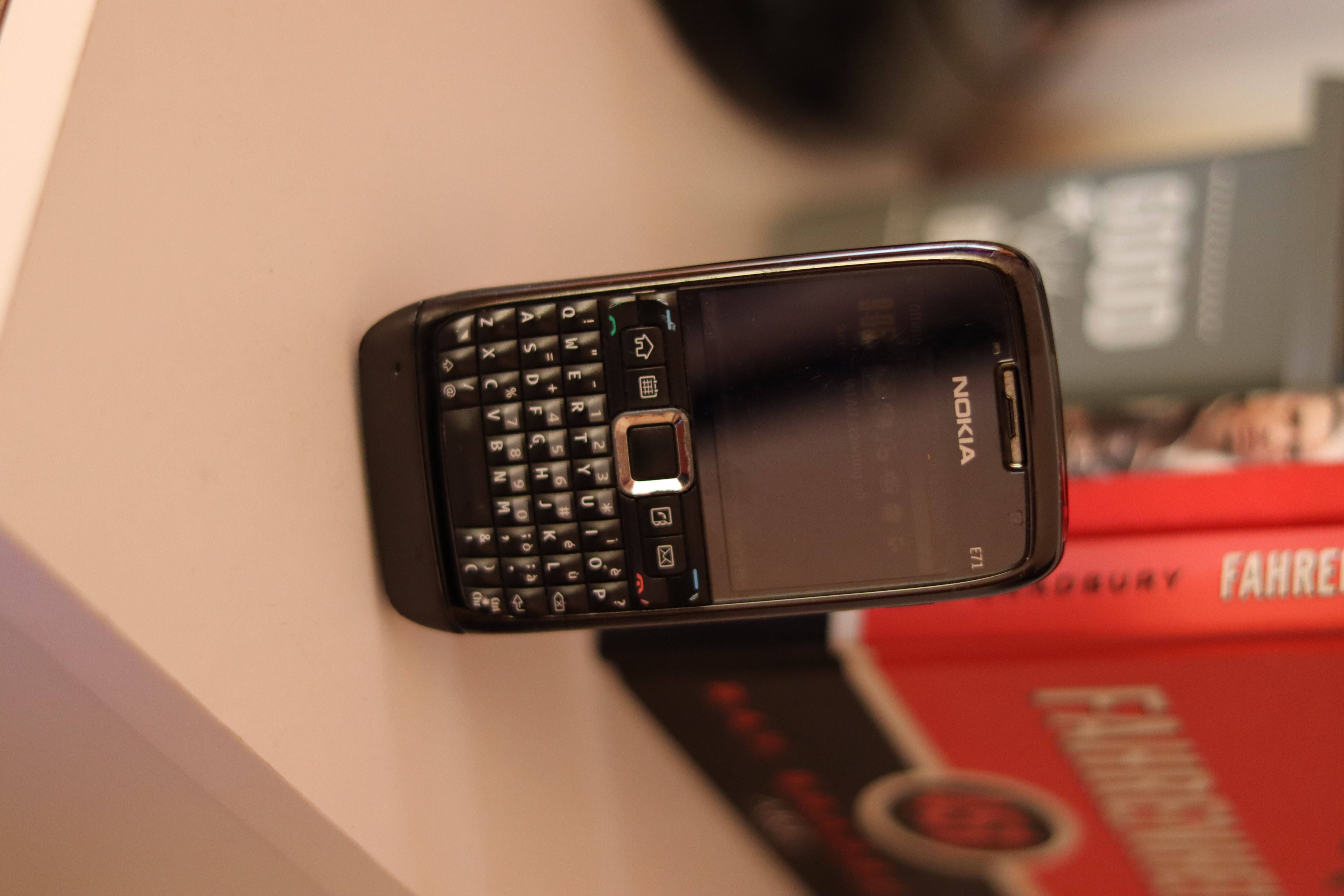 諾基亞 桌布 壁紙 — 一部黑色Nokia E71智慧型手機配備實體鍵盤，放在白色表面上，旁邊有紅色書籍。