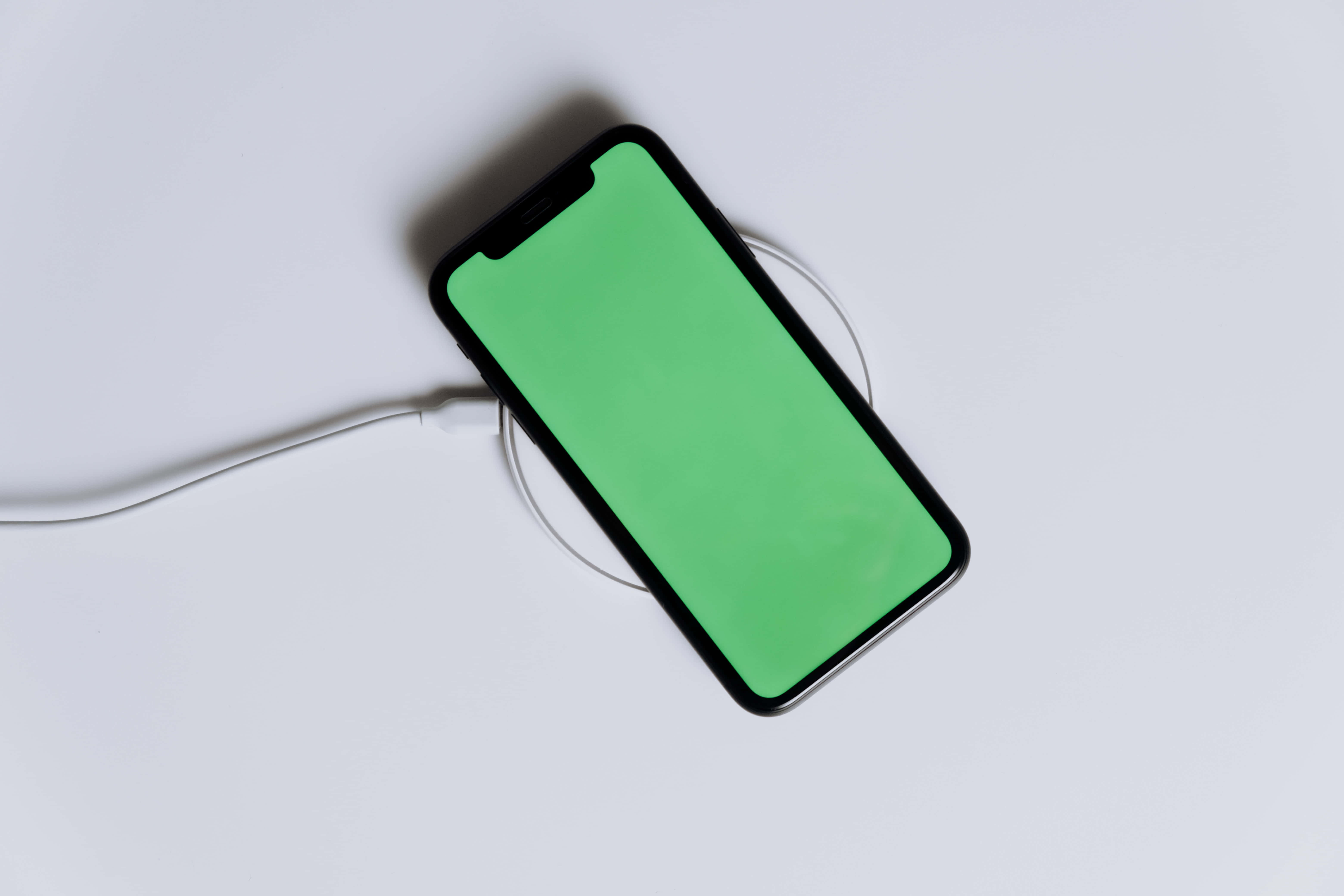 iPhone 桌布 壁紙 — 一部黑色智慧型手機帶有綠色螢幕，放在一個白色充電板上，由一條白色電線連接，置於純白色表面上。