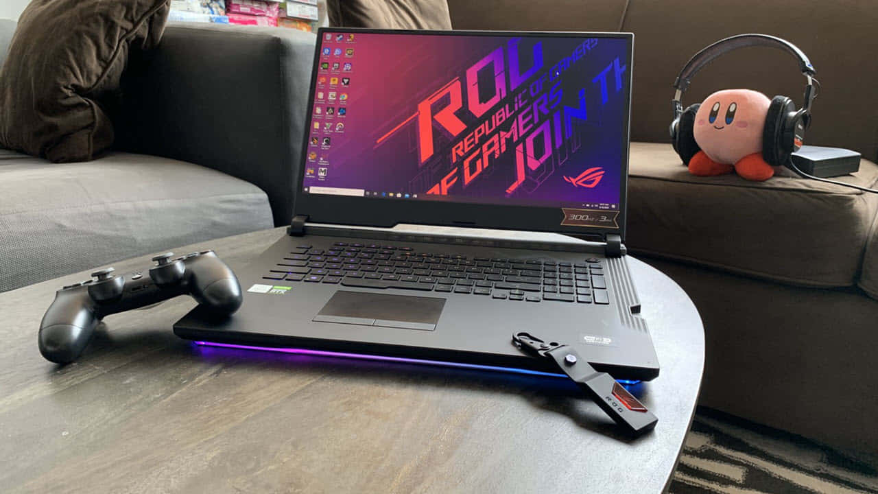 華碩 ROG 桌布 壁紙 — 一部黑色遊戲筆記型電腦帶有紫色LED光芒，放在木桌上，旁邊有一個控制器和戴著耳機的卡比絨毛玩具。