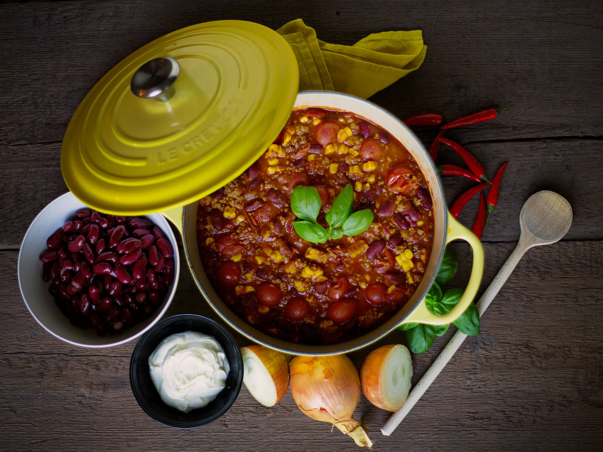 Mexican Food 桌布 壁紙 — 一鍋冒著熱氣的濃郁辣椒，配上羅勒點綴，放在深色木桌上，周圍是新鮮食材和木勺。