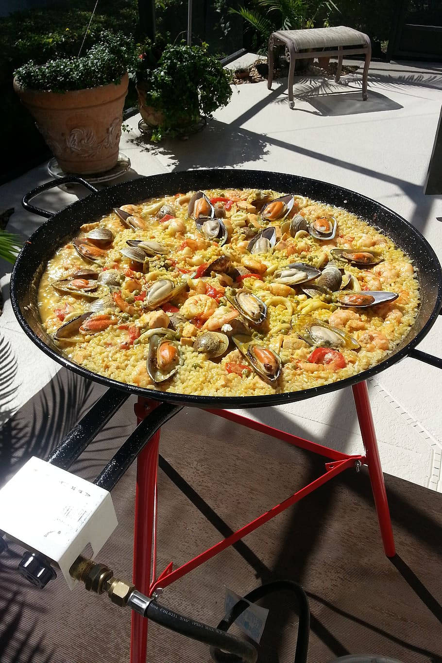 烹飪 桌布 壁紙 — 一鍋色彩繽紛的海鮮西班牙大鍋飯在黑色平底鍋中滋滋作響，放在紅色支架上，位於陽光普照的露台下。