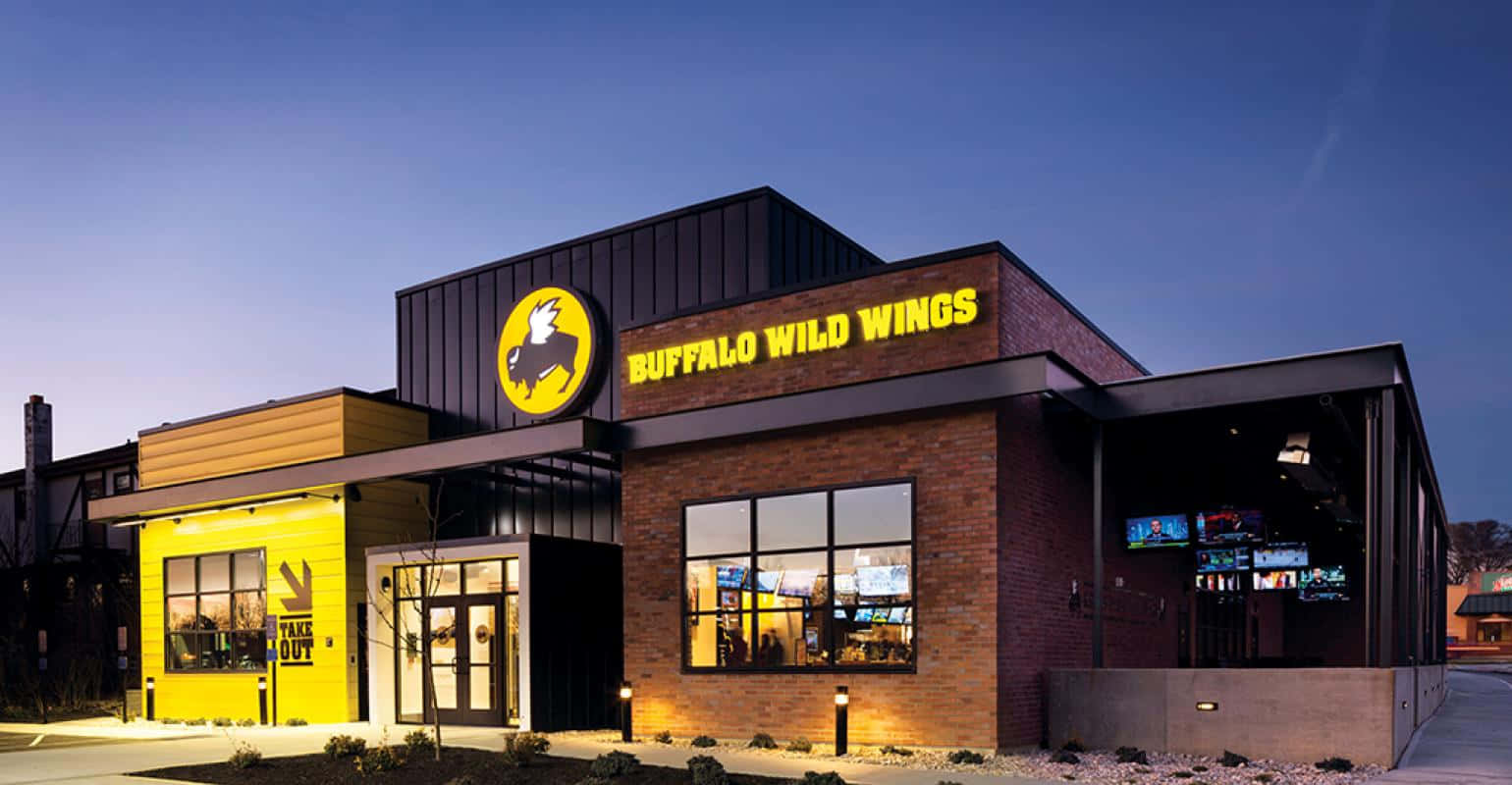 水牛城辣雞翅 桌布 壁紙 — 一間 Buffalo Wild Wings 餐廳在黃昏時分亮起，帶有發光的黃色招牌和溫暖的室內燈光，襯托著深藍色的天空。
