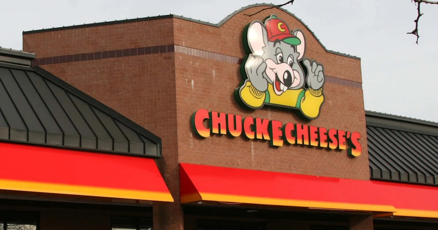 Chuck E Cheese 桌布 壁紙 — 一間 Chuck E Cheese's 餐廳，卡通老鼠吉祥物在磚造建築上於多雲天空下揮手。