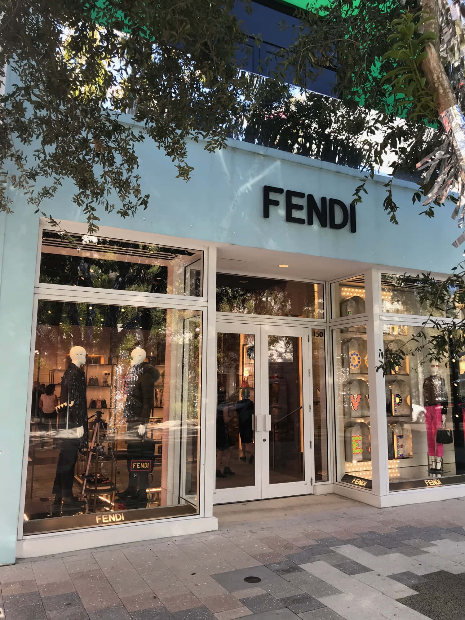 Fendi 桌布 壁紙 — 一間FENDI豪華精品店，淺藍色外牆和玻璃櫥窗，展示人體模型，上方樹枝投下斑駁陰影。