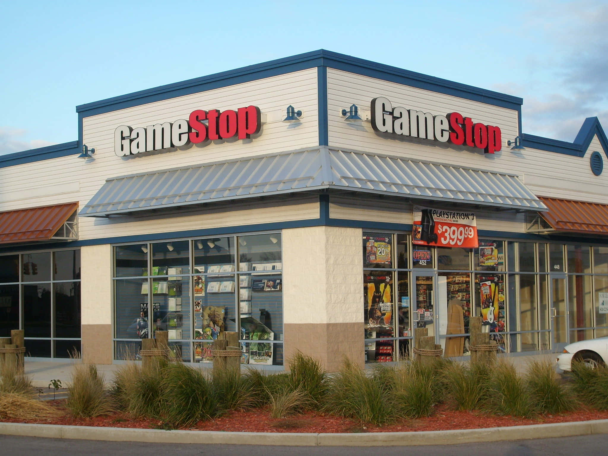 GameStop 桌布 壁紙 — 一間GameStop商店，帶有紅色和白色的招牌，以及大型玻璃櫥窗，在晴朗的藍天下展示電子遊戲和主機。