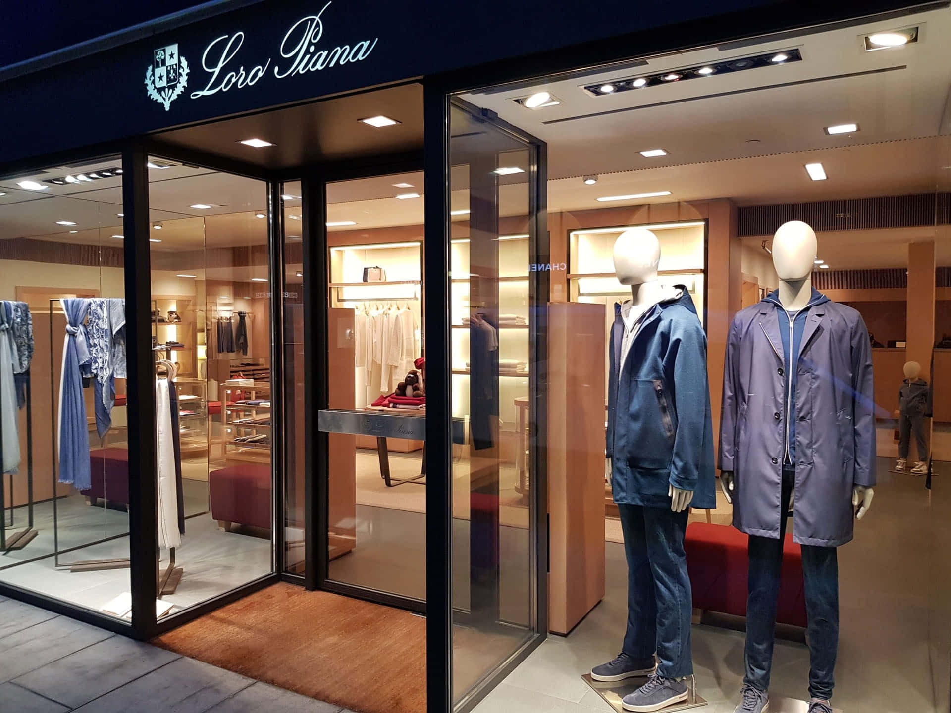Mens Fashion 桌布 壁紙 — 一間Loro Piana奢華男裝店，玻璃櫥窗內展示穿著藍色外套和牛仔褲的人體模型，在溫暖的室內燈光下。
