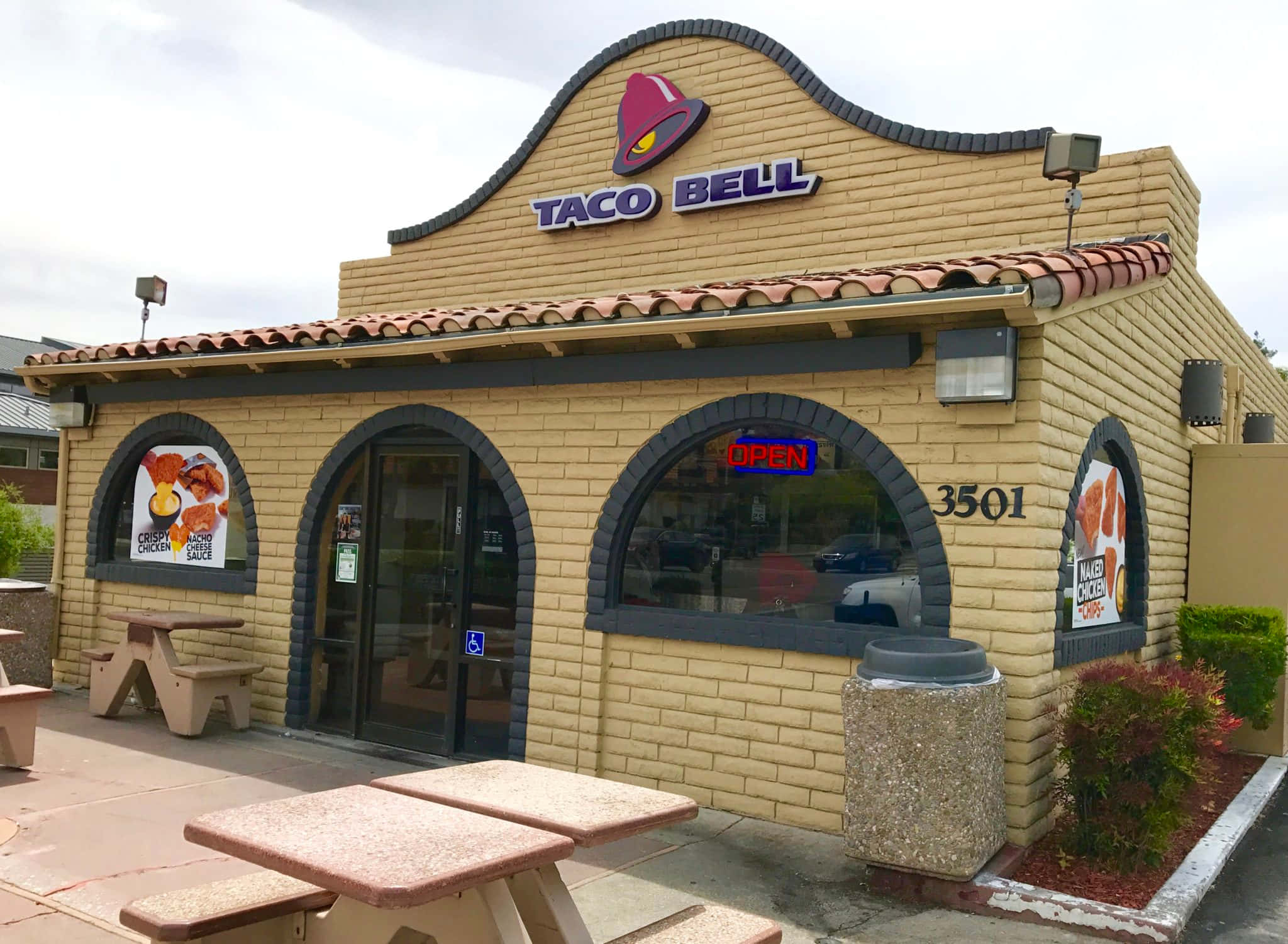 塔可鐘 桌布 壁紙 — 一間Taco Bell餐廳，有米色磚牆、拱形窗戶和野餐桌，天空多雲。
