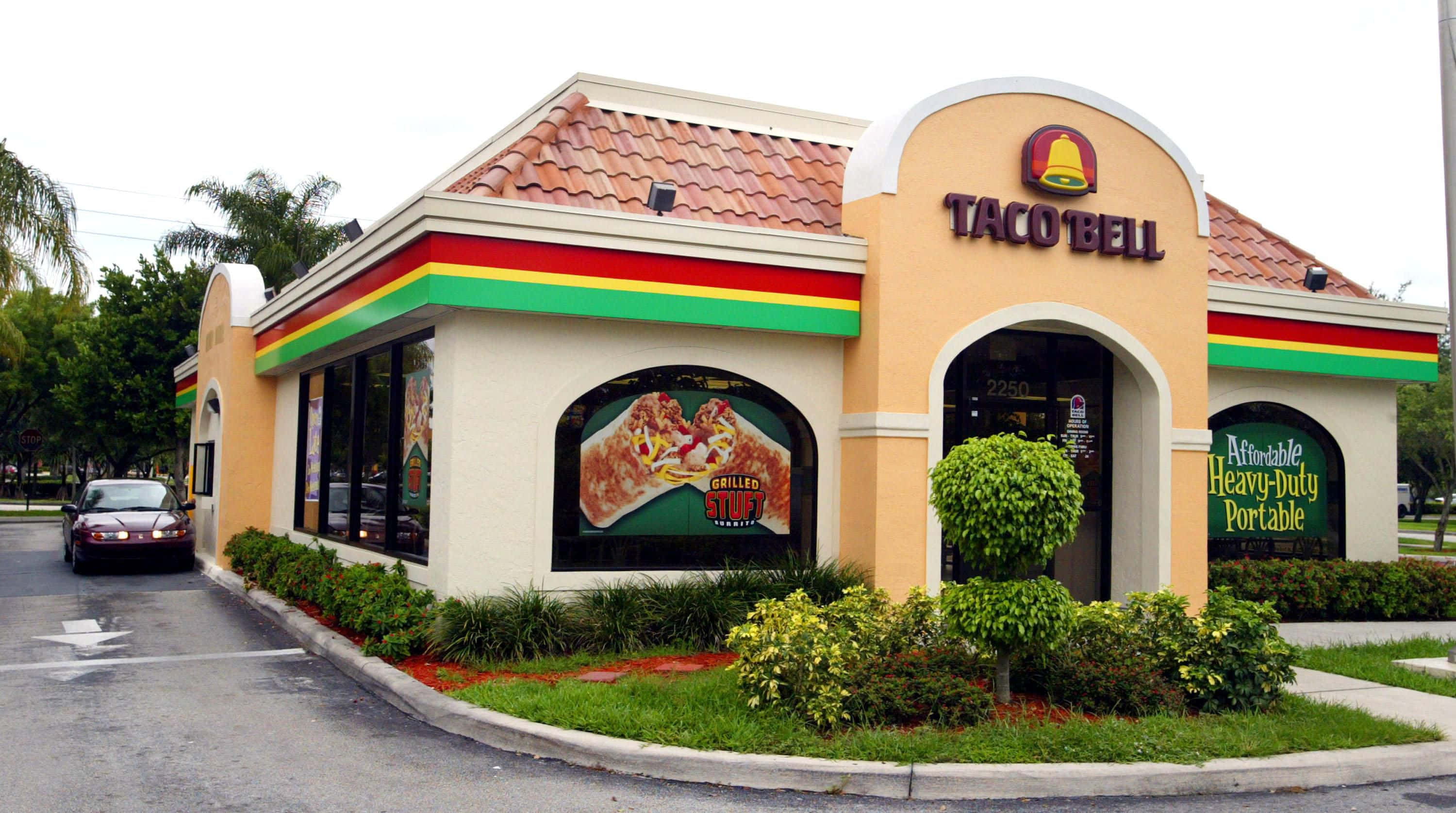 塔可鐘 桌布 壁紙 — 一間Taco Bell餐廳，米色灰泥外牆和紅色瓦片屋頂，周圍環繞著綠色景觀和棕櫚樹。