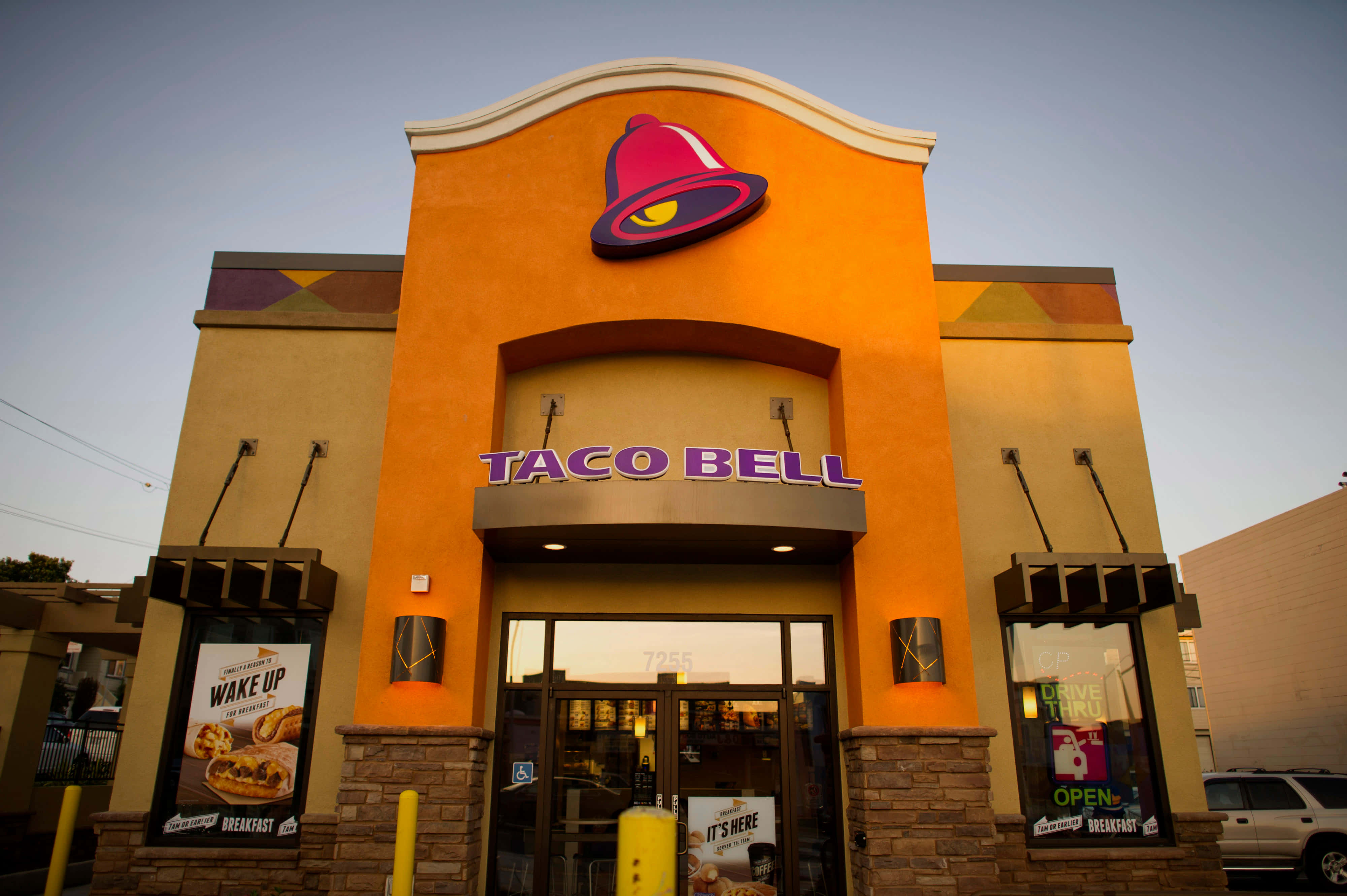 Taco Bell 桌布 壁紙 — 一間Taco Bell餐廳在黃昏時分，溫暖的橙色牆壁和發光的紫色招牌，襯托著柔和的藍天。