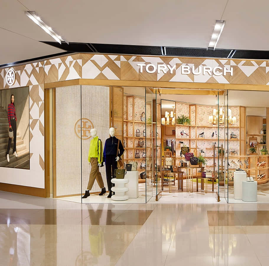 Tory Burch 桌布 壁紙 — 一間Tory Burch精品店以玻璃牆展示穿著鮮豔服裝的人體模型和陳列手袋的貨架，在溫暖的燈光下。