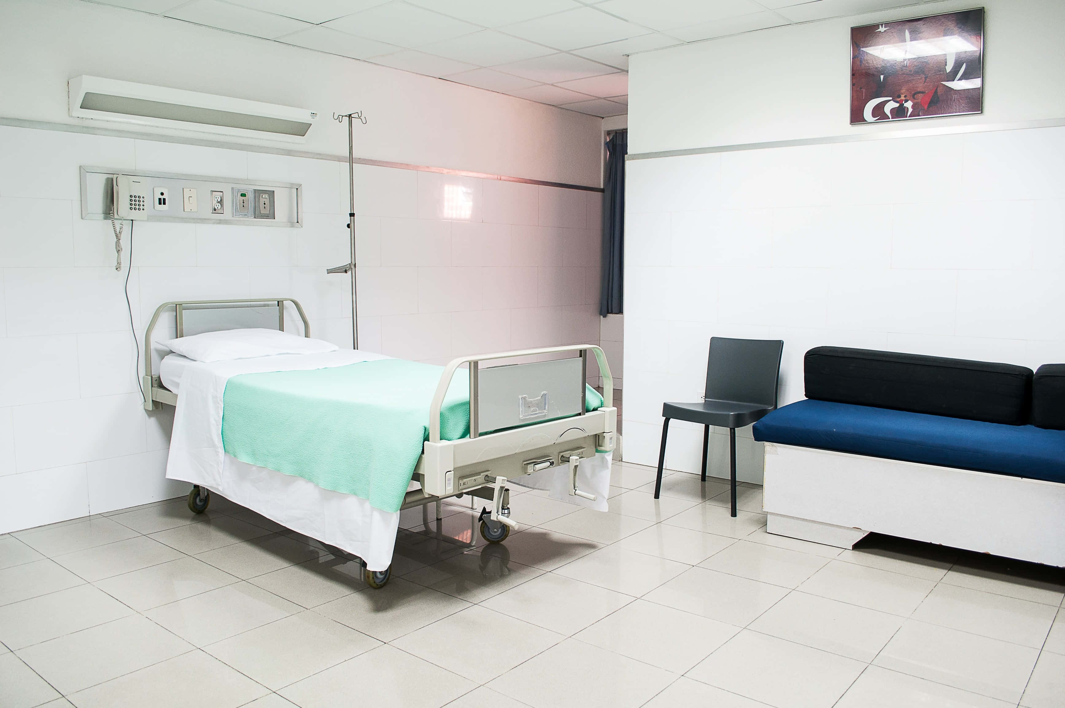 Hospital 桌布 壁紙 — 一間乾淨的醫院病房，內有鋪著薄荷綠色床單的醫療床，旁邊有一張藍色沙發，在螢光燈下。