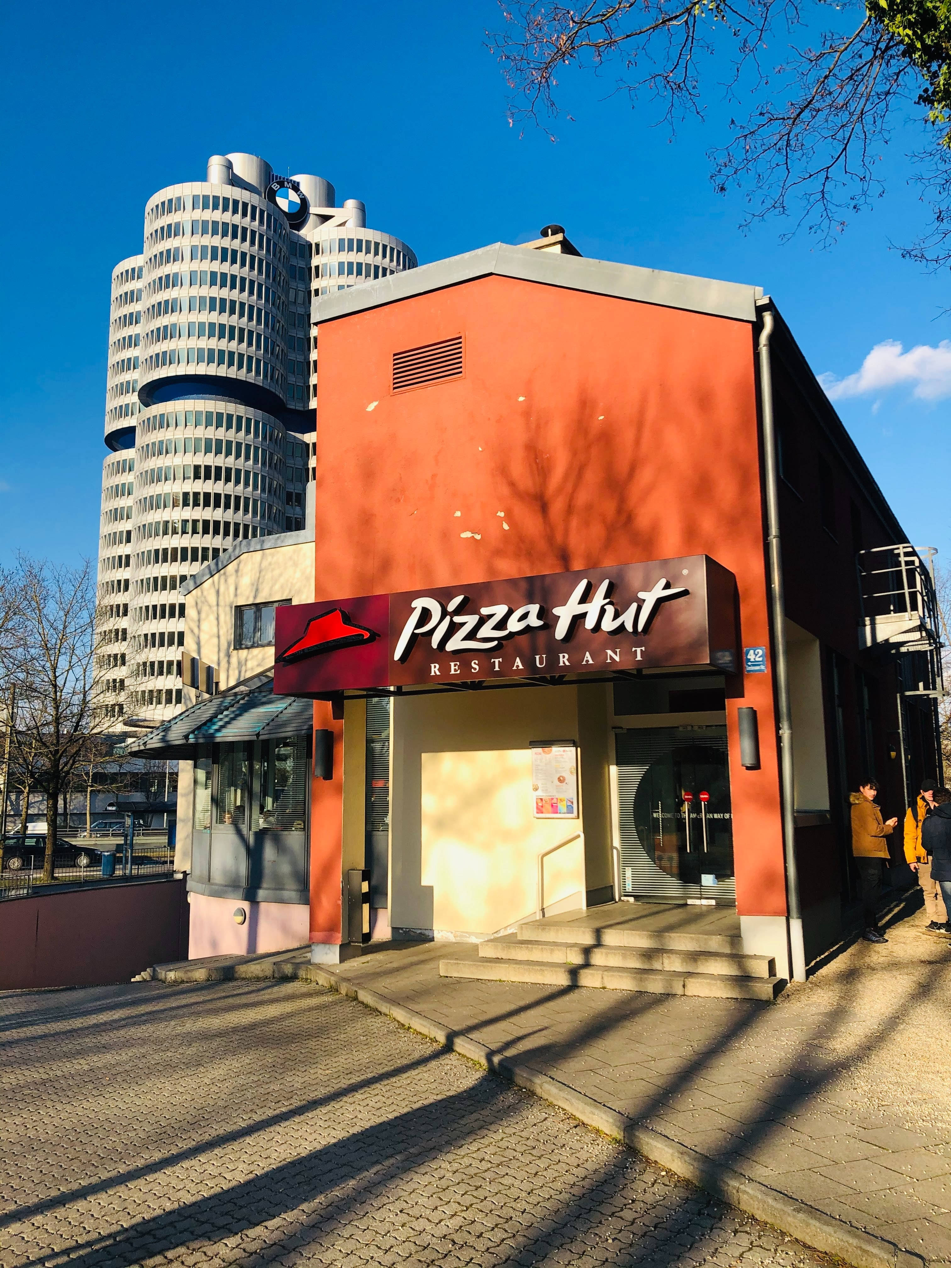Hut 桌布 壁紙 — 一間亮橙色的必勝客餐廳矗立在標誌性的BMW總部大樓前，天空湛藍清澈。