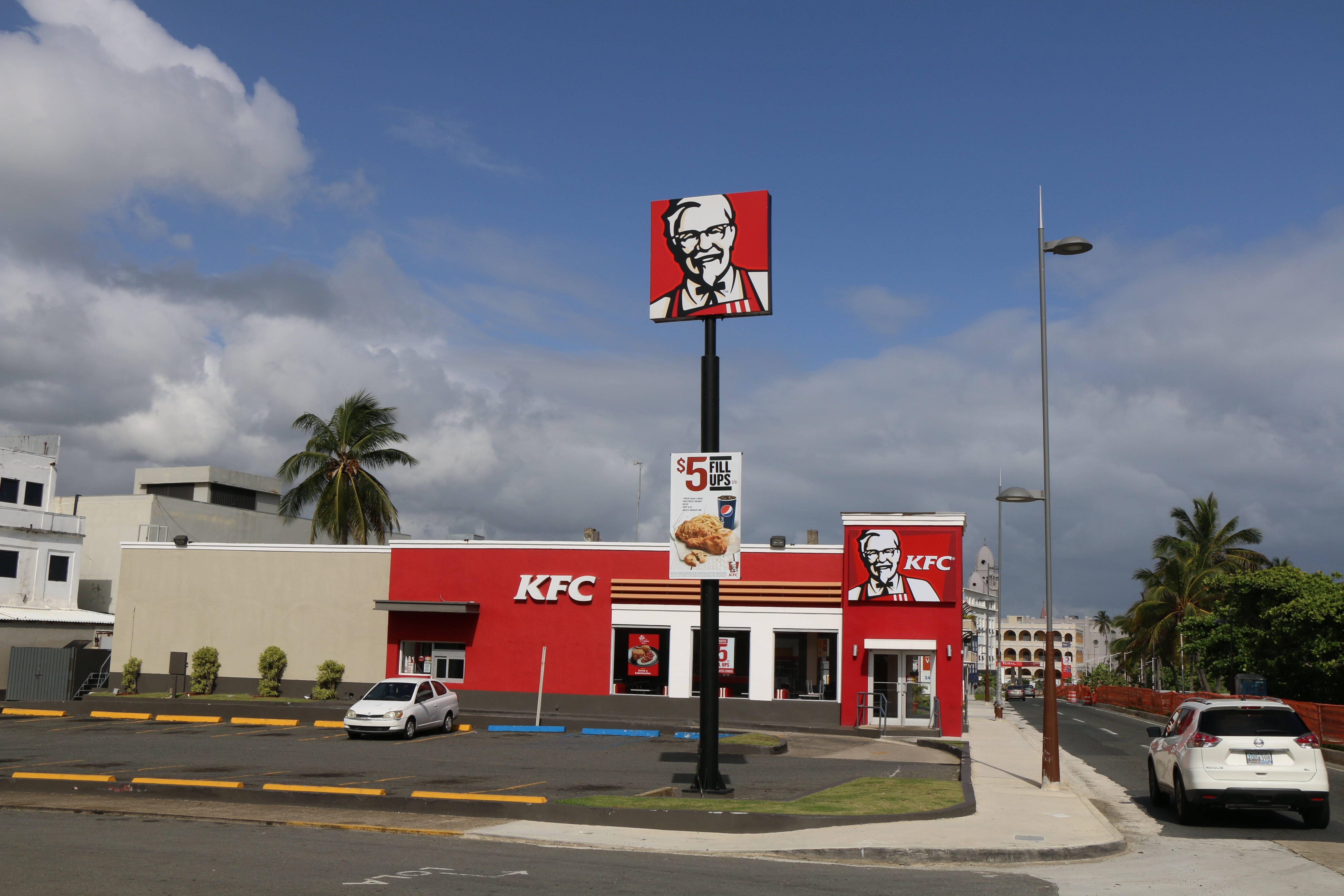 Kfc 桌布 壁紙 — 一間亮紅色的KFC餐廳，天空部分多雲，有棕櫚樹和停車場的汽車。