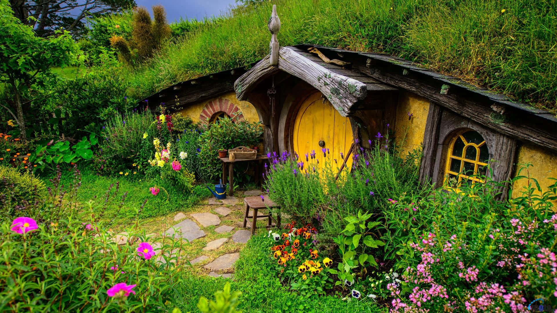 Picturesque 桌布 壁紙 — 一間充滿奇思妙想的哈比人小屋，黃色大門搭配綠色屋頂，坐落於色彩繽紛、生機盎然的花園中。