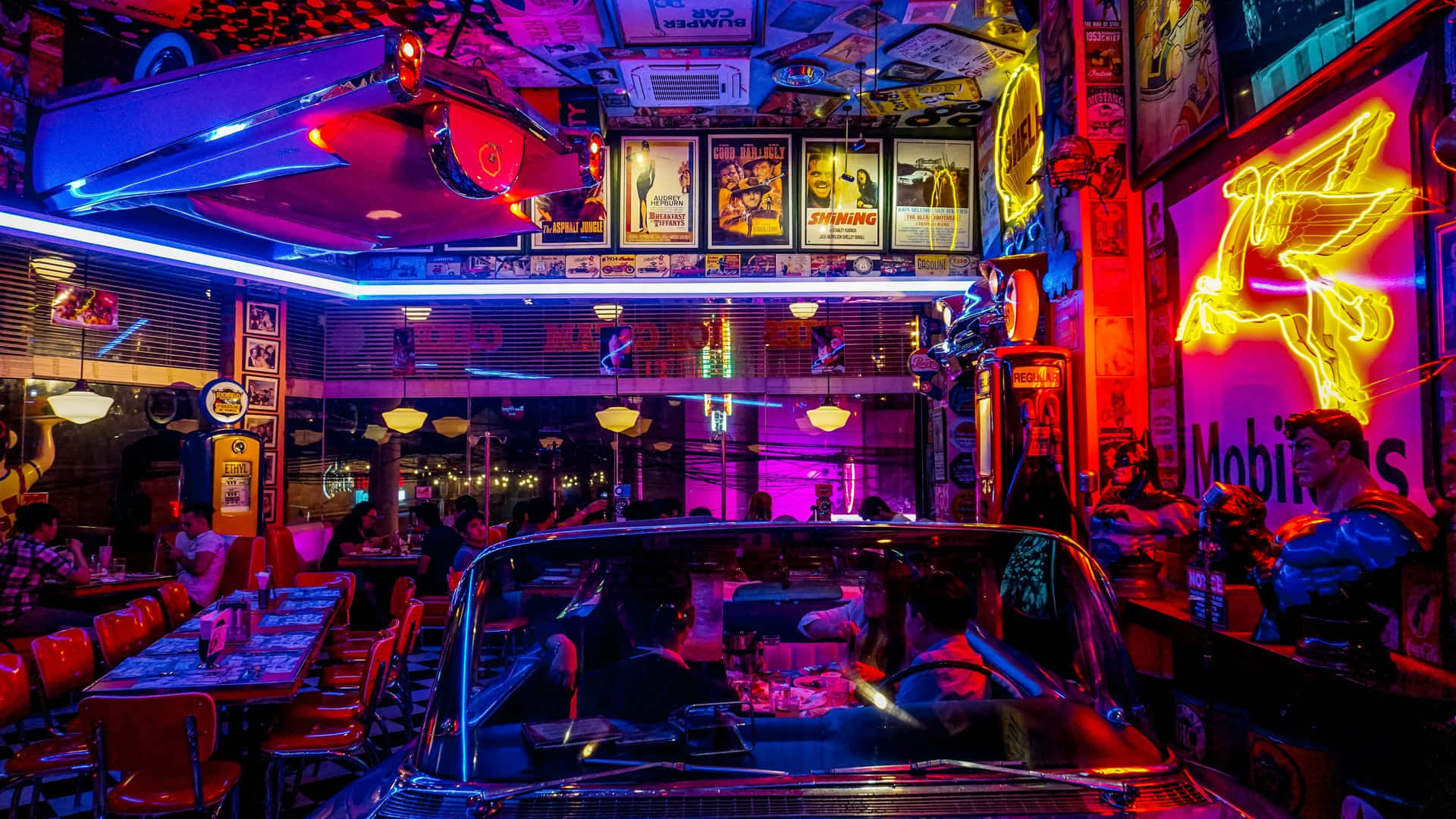 club 桌布 壁紙 — 一間充滿霓虹燈光的復古餐廳，內裡停著一輛經典汽車，顧客在鮮豔的粉紅和藍色光芒下用餐。