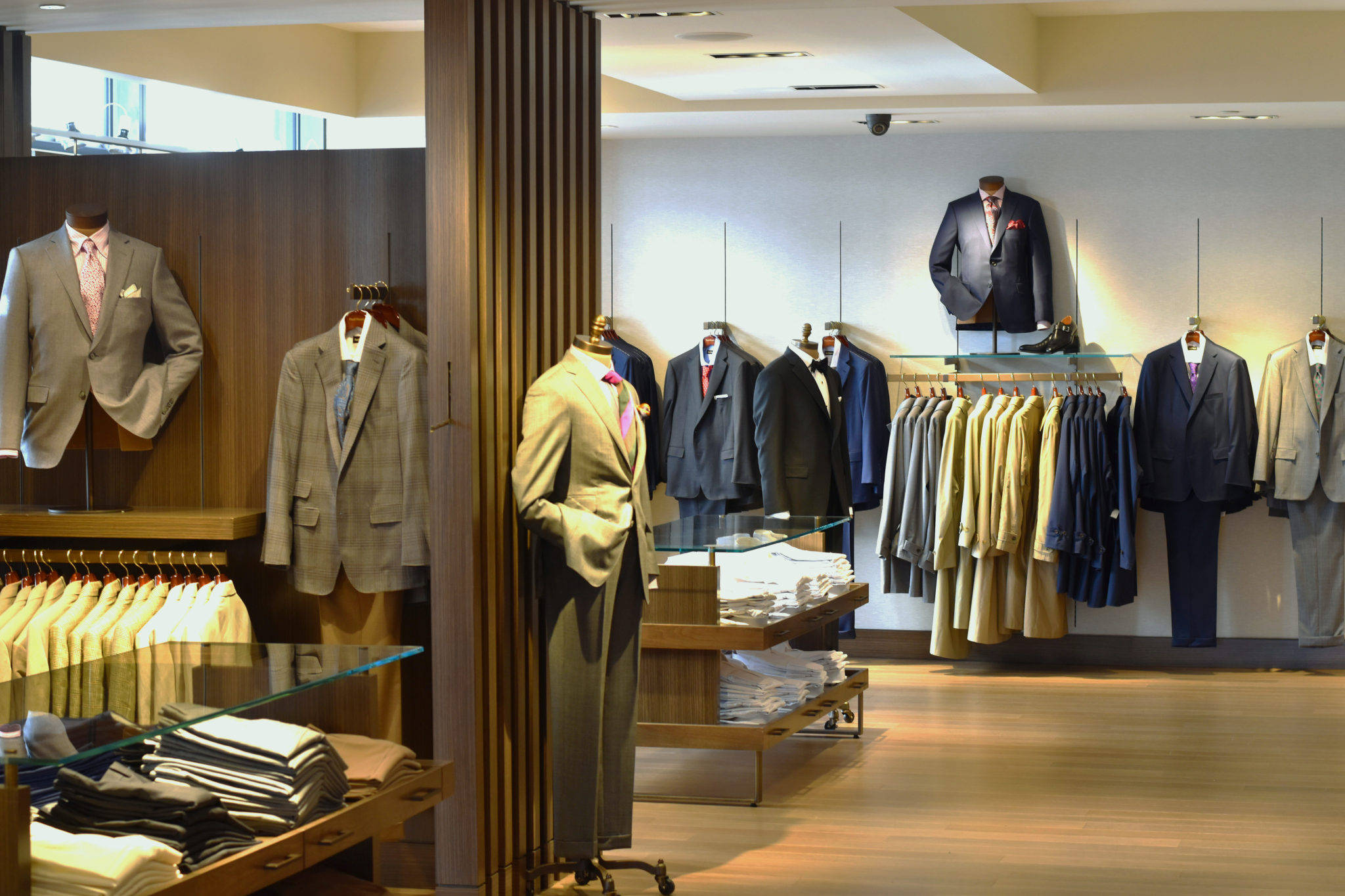 Clothes 桌布 壁紙 — 一間光線充足的高級男裝精品店，在模特兒和衣架上展示訂製西裝，搭配溫暖的木質裝飾和玻璃層架。