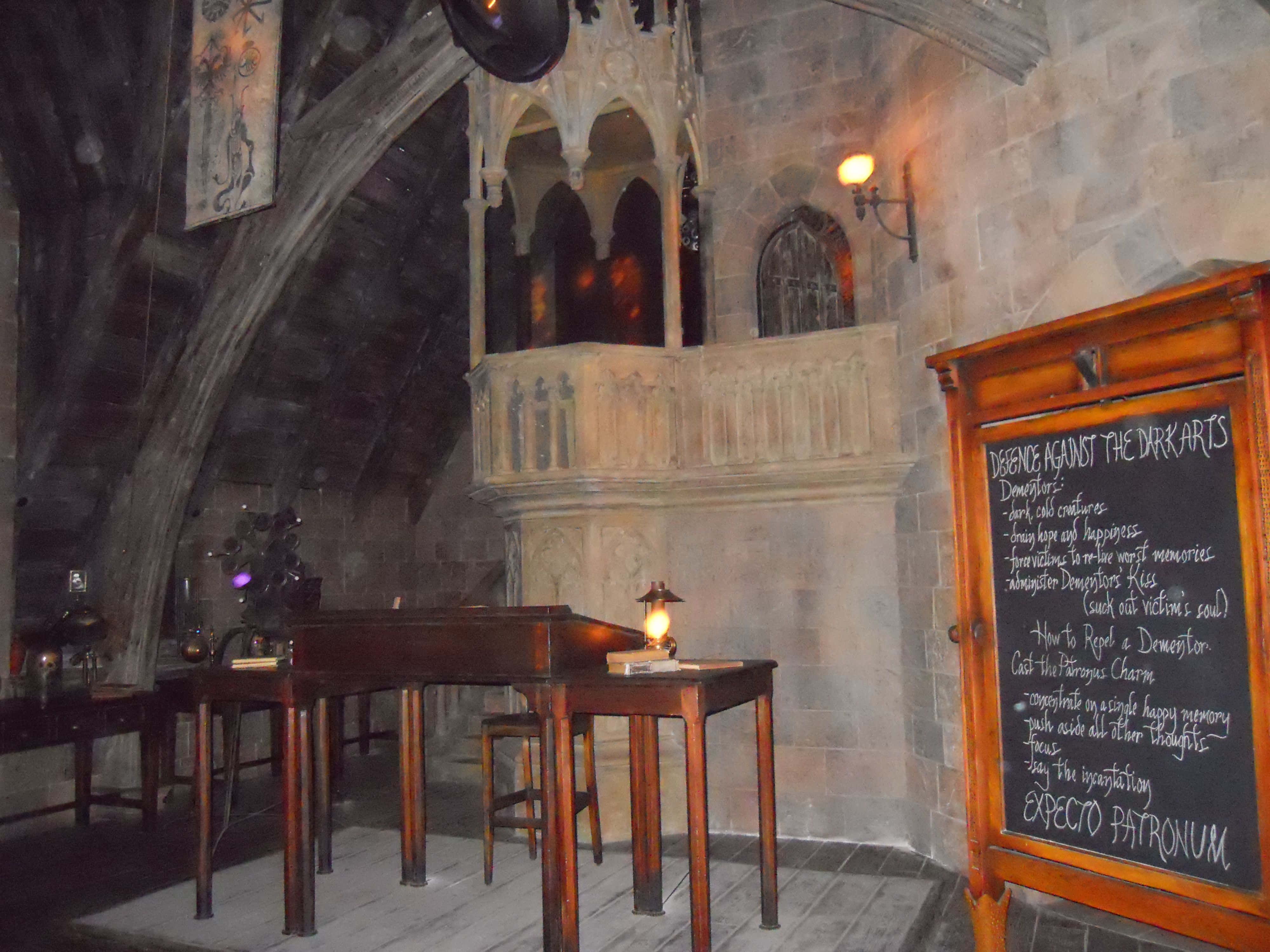 The Hogwarts Dark Arts Classroom 桌布 壁紙 — 一間光線昏暗的石頭教室，配有木製書桌和列出黑魔法防禦術咒語的黑板。