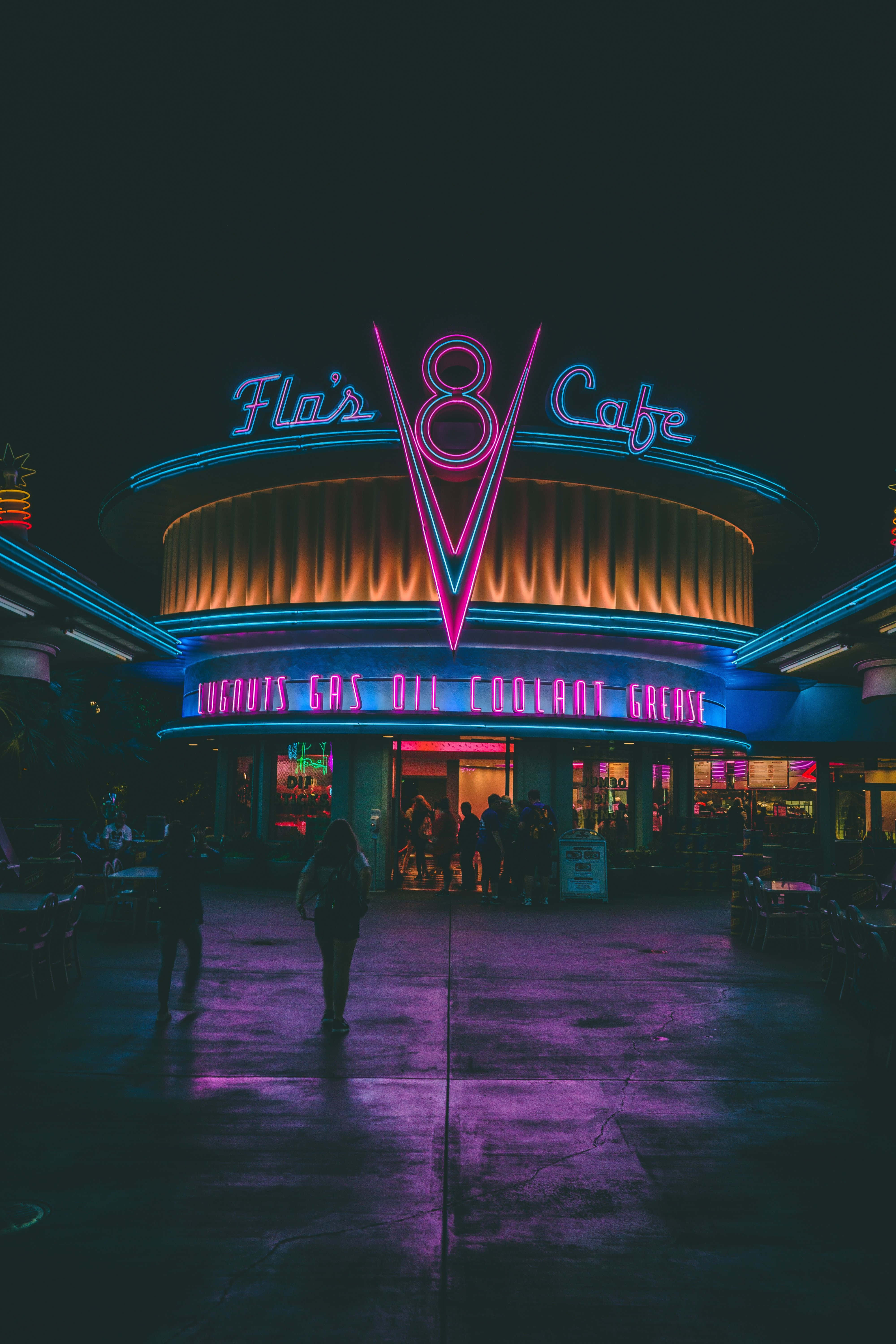 iPhone Neon 桌布 壁紙 — 一間名為 Fla's V8 Cafe 的復古餐廳在漆黑的夜空下閃爍著粉紅和藍色的霓虹燈光，剪影人物正走向入口。