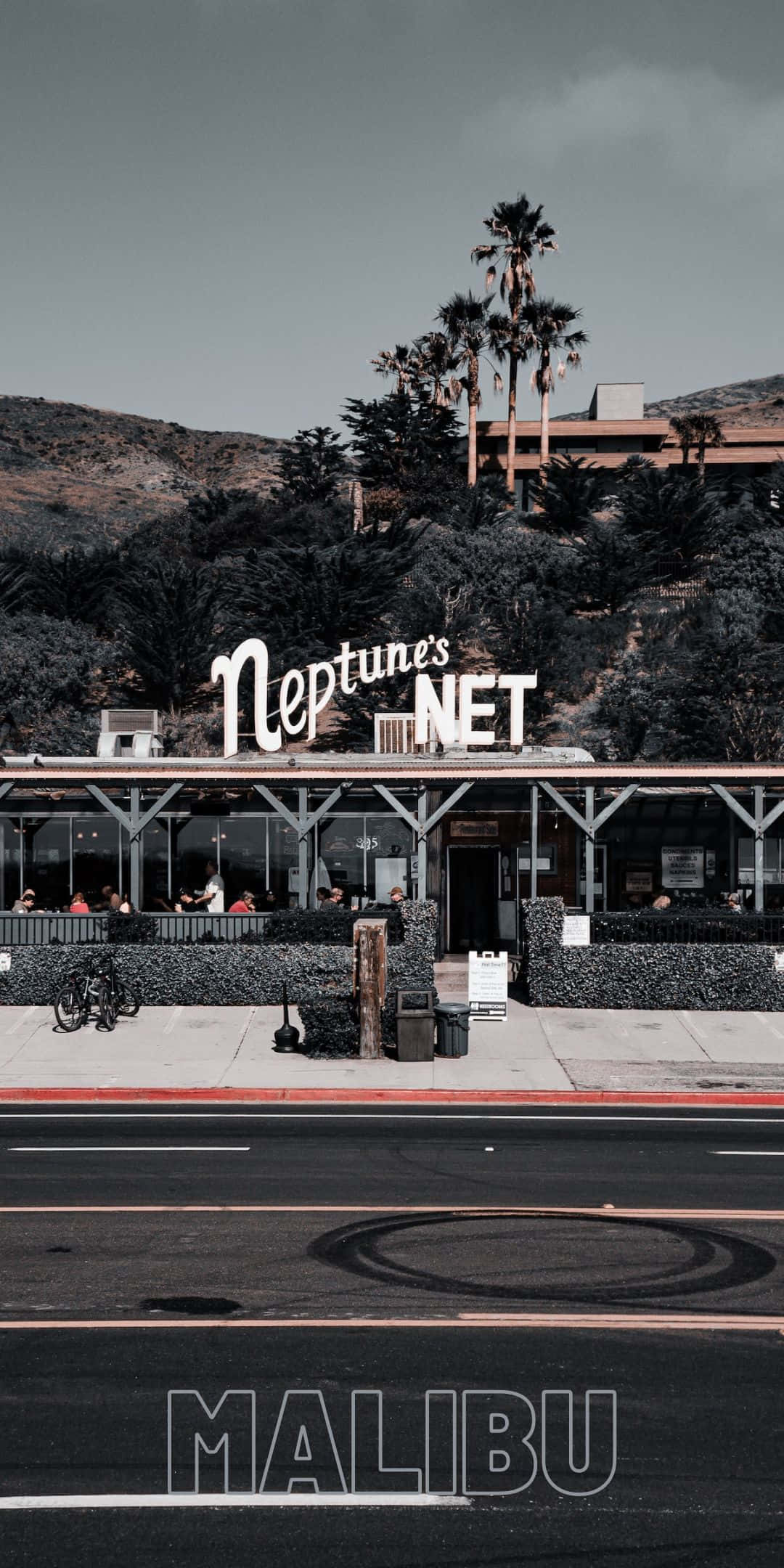 加州 桌布 壁紙 — 一間名為 Neptune's Net 的復古餐廳位於沿海街道上，背後有棕櫚樹和山丘，天空陰沉。