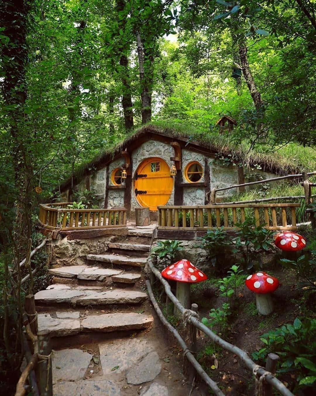 cabin 桌布 壁紙 — 一間奇幻風格的哈比人式石屋，擁有長滿青苔的屋頂和明亮的黃色大門，坐落在鬱鬱蔥蔥的綠色森林中。