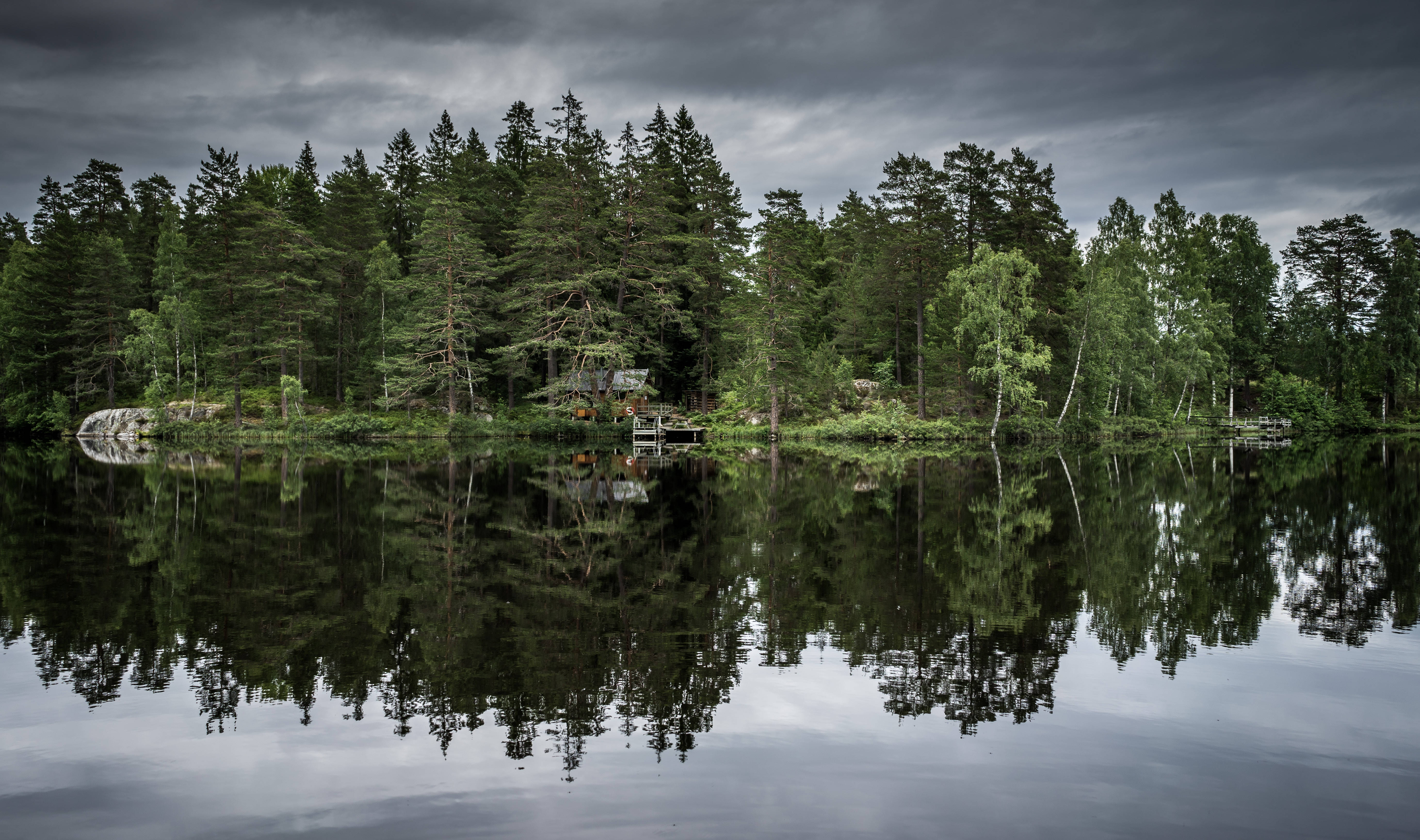 Lake 桌布 壁紙 — 一間孤獨的小屋坐落在茂密的松樹林中，位於平靜的湖面上，在沉重的灰色天空下呈現完美的鏡面倒影。