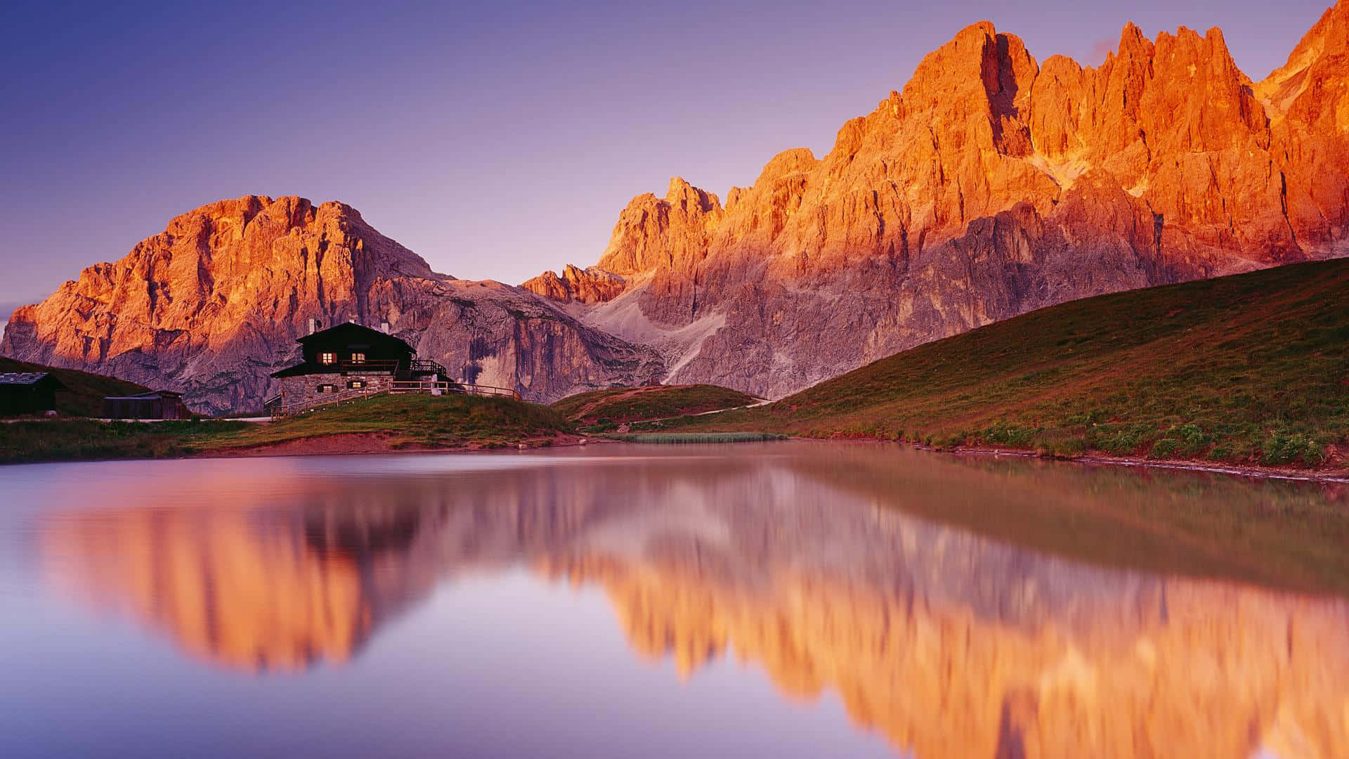 National Parks 桌布 壁紙 — 一間孤獨的山間小屋倒映在平靜的水面上，金色的山峰在日落時分閃耀。