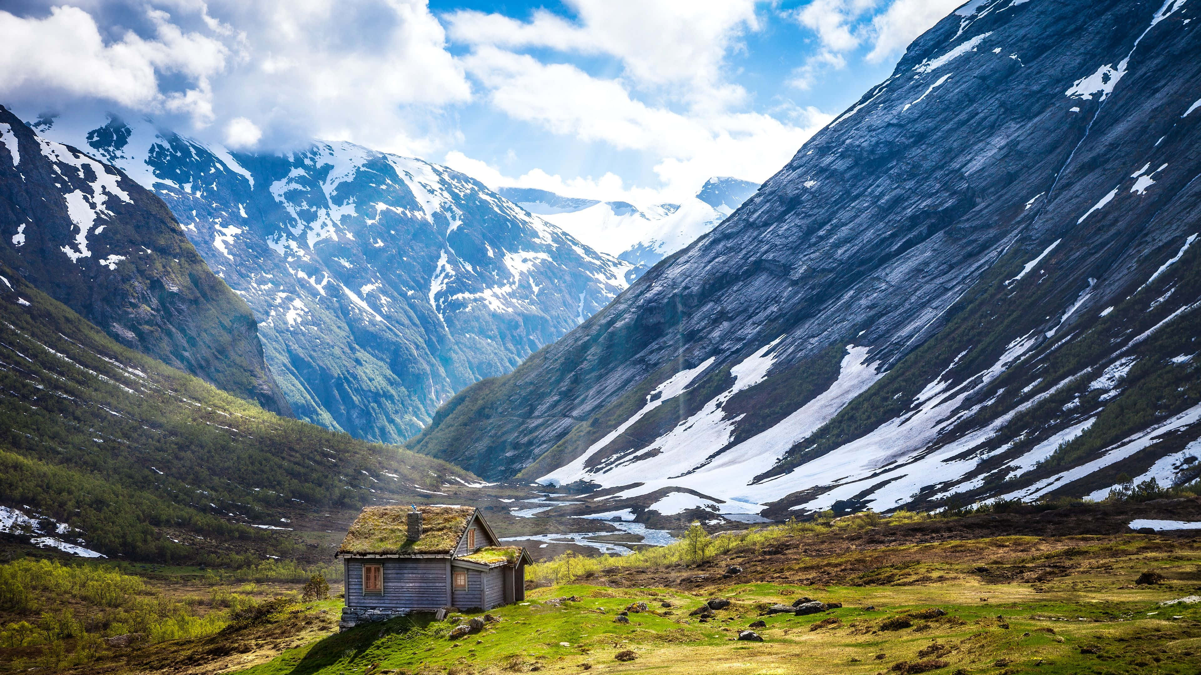 Norway 桌布 壁紙 — 一間孤獨的木屋，屋頂長滿苔蘚，坐落在陽光明媚的山谷中，周圍是白雪覆蓋的高聳山脈，頭頂是明亮的藍天和蓬鬆的白雲。