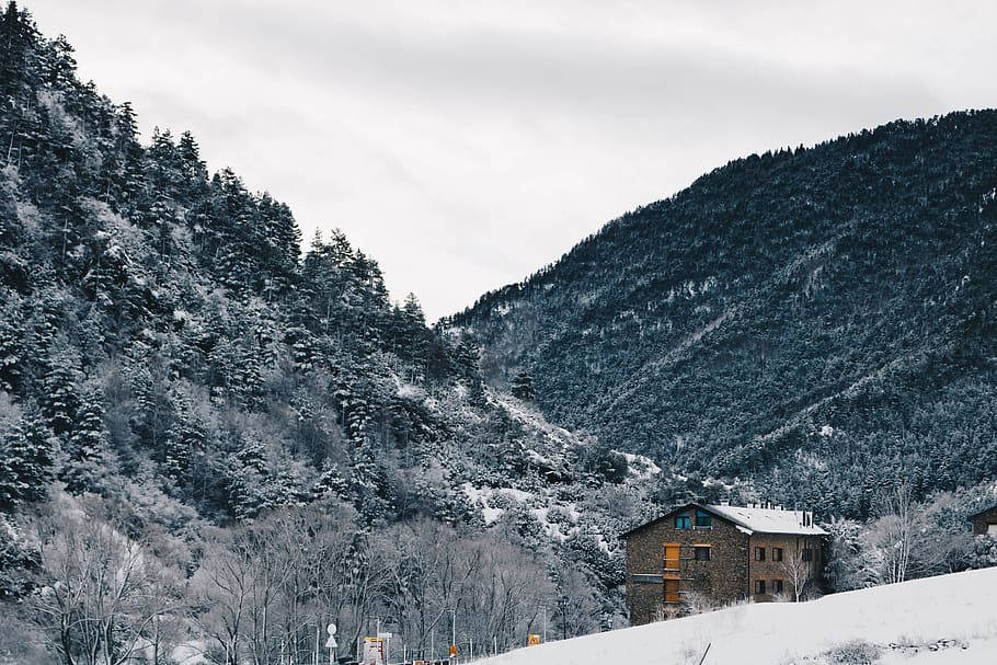 山景 桌布 壁紙 — 一間孤獨的石屋坐落在白雪覆蓋的山谷中，四周環繞著常綠森林和巍峨的山脈，天空陰沉。
