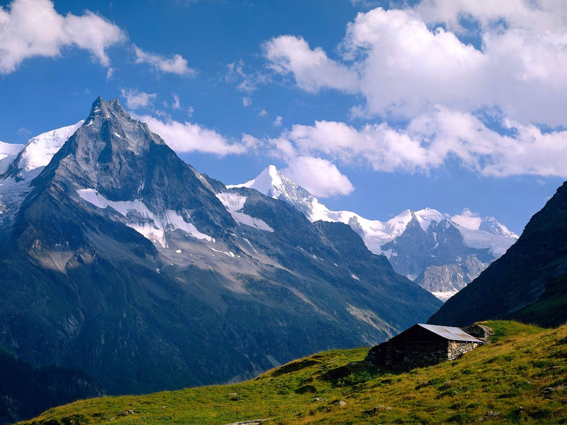 Alps 桌布 壁紙 — 一間孤獨的石屋坐落於陽光明媚的草地上，下方是白雪覆蓋的山峰，上方是明亮的藍天和蓬鬆的白雲。