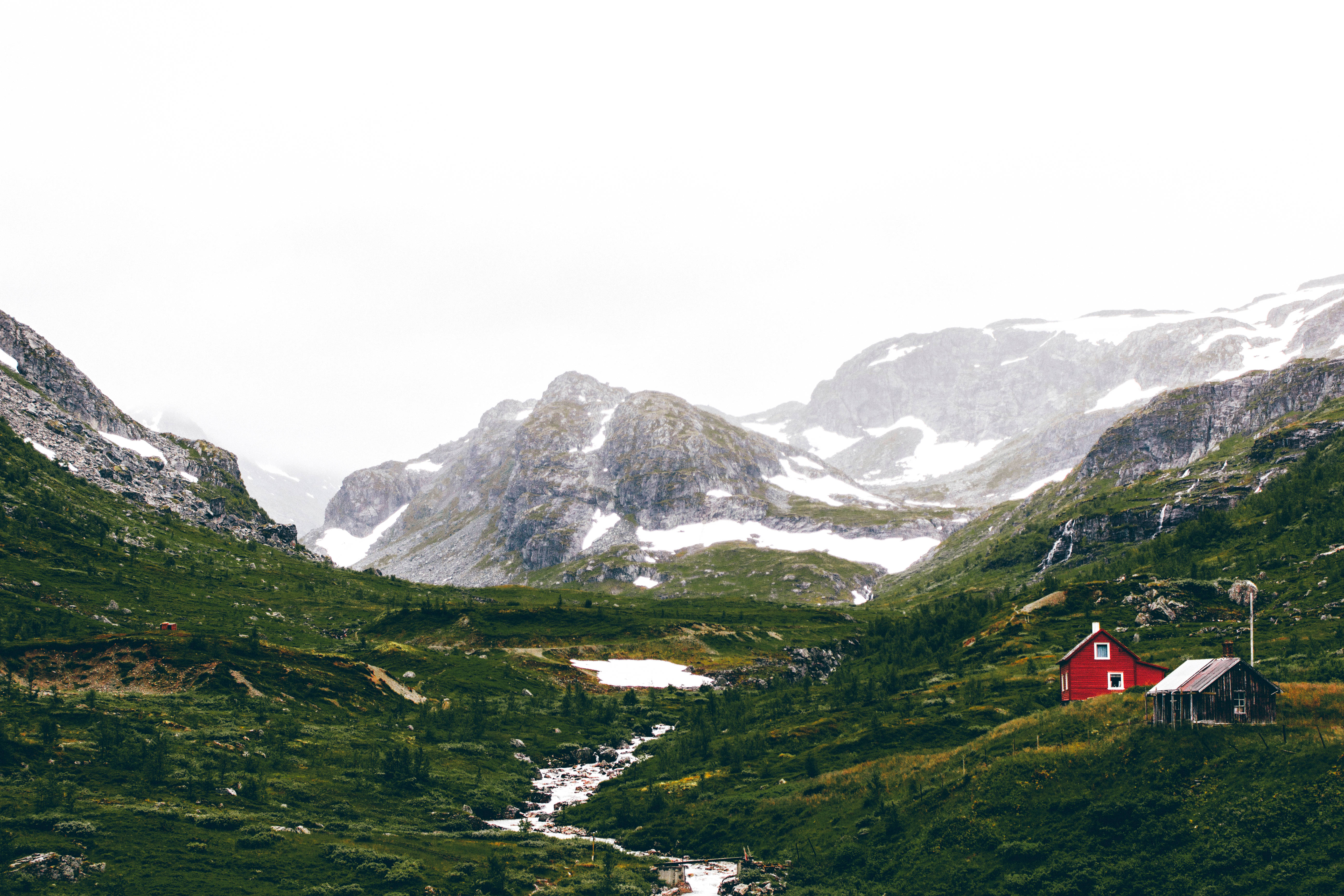 Norway 桌布 壁紙 — 一間孤獨的紅色小屋坐落在鬱鬱蔥蔥的綠色山谷中，白雪覆蓋的山脈在陰天天空下聳立。