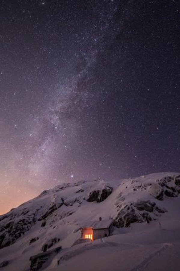iOS 8 桌布 壁紙 — 一間孤獨的雪覆蓋小屋在壯麗的星空下溫暖地發光，銀河拱在頭頂。