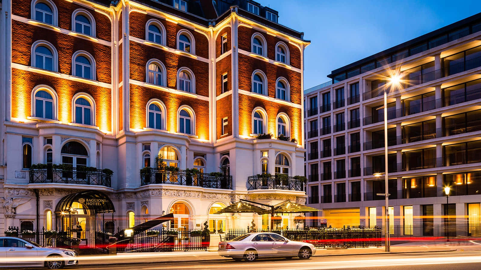 Hotel 桌布 壁紙 — 一間宏偉的磚造酒店散發著溫暖的金色燈光，在暮色天空下閃耀，汽車在繁忙的城市街道上疾馳而過。