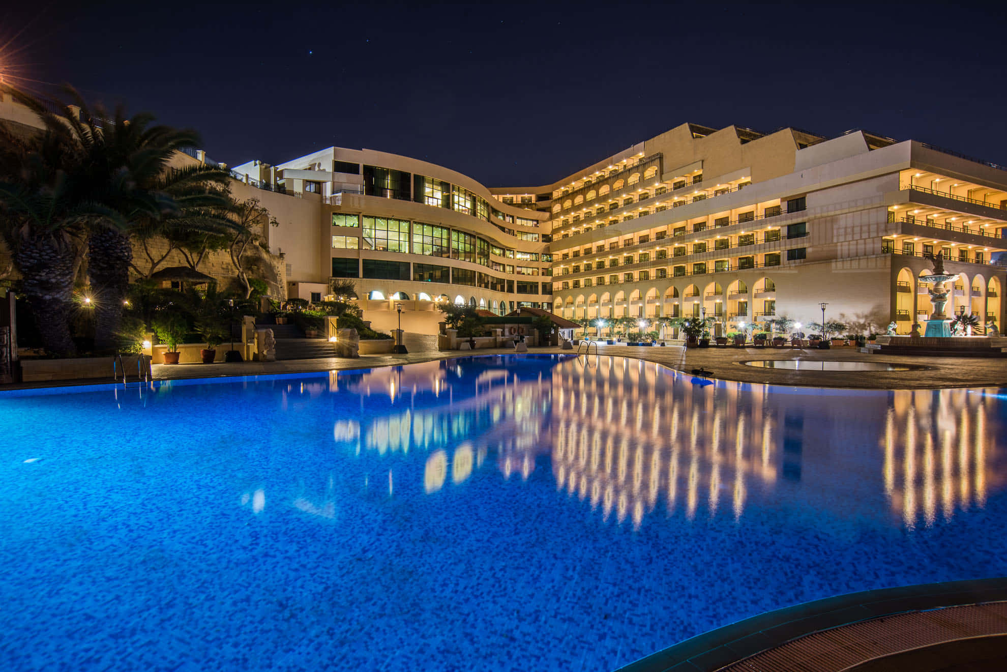 Hotel 桌布 壁紙 — 一間宏偉的酒店在夜晚溫暖地發光，其燈光完美地映照在星空下廣闊的藍色泳池中。