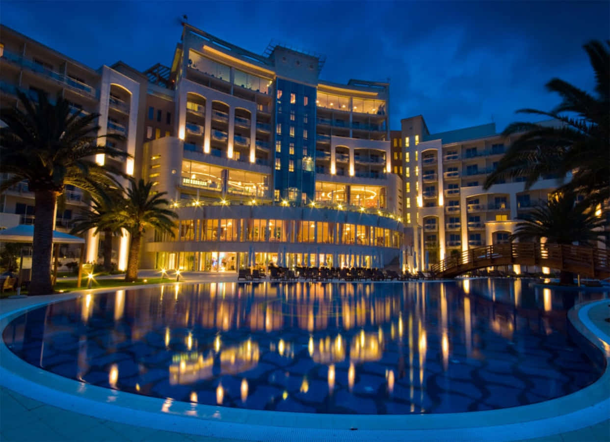 Hotel 桌布 壁紙 — 一間宏偉的酒店在黃昏時亮起燈光，溫暖的燈光倒映在棕櫚樹環繞的大型泳池中。