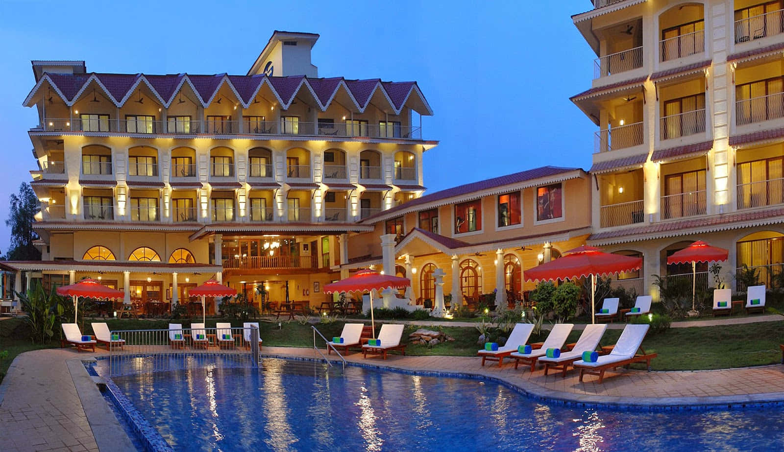 Hotel 桌布 壁紙 — 一間宏偉的飯店，溫暖的燈光和紅色遮陽傘環繞著暮色中寧靜的泳池。