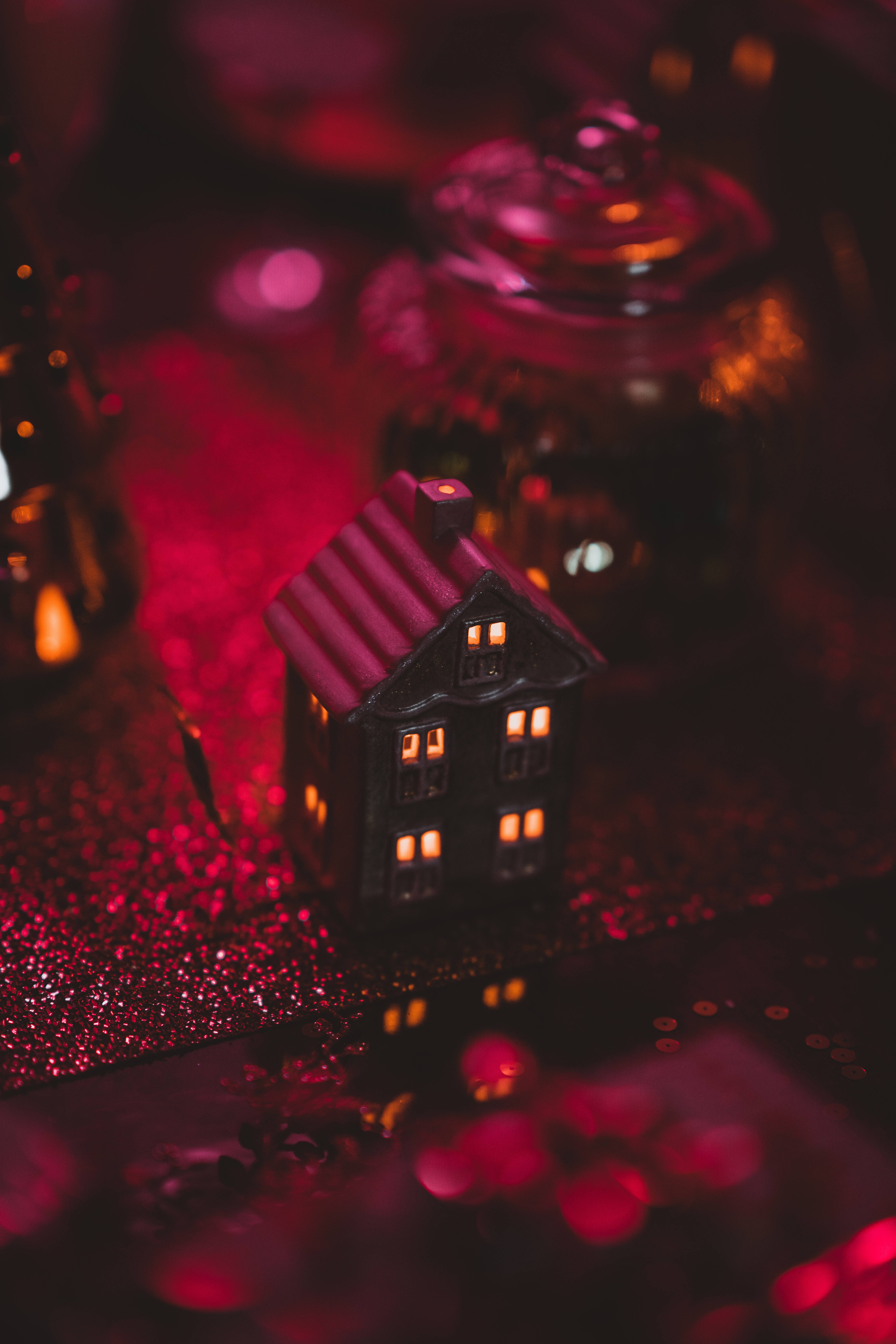 lights 桌布 壁紙 — 一間帶有發光窗戶的微型黑色房屋坐在閃閃發光的紅色表面上，周圍環繞著模糊的節日玻璃罐。