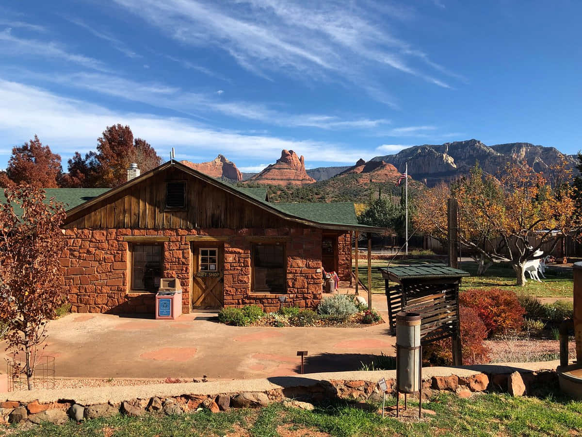 Sedona 桌布 壁紙 — 一間帶木質裝飾的質樸石屋坐落於明亮的藍天下，被秋天的樹木和遠處的紅色岩層所環繞。