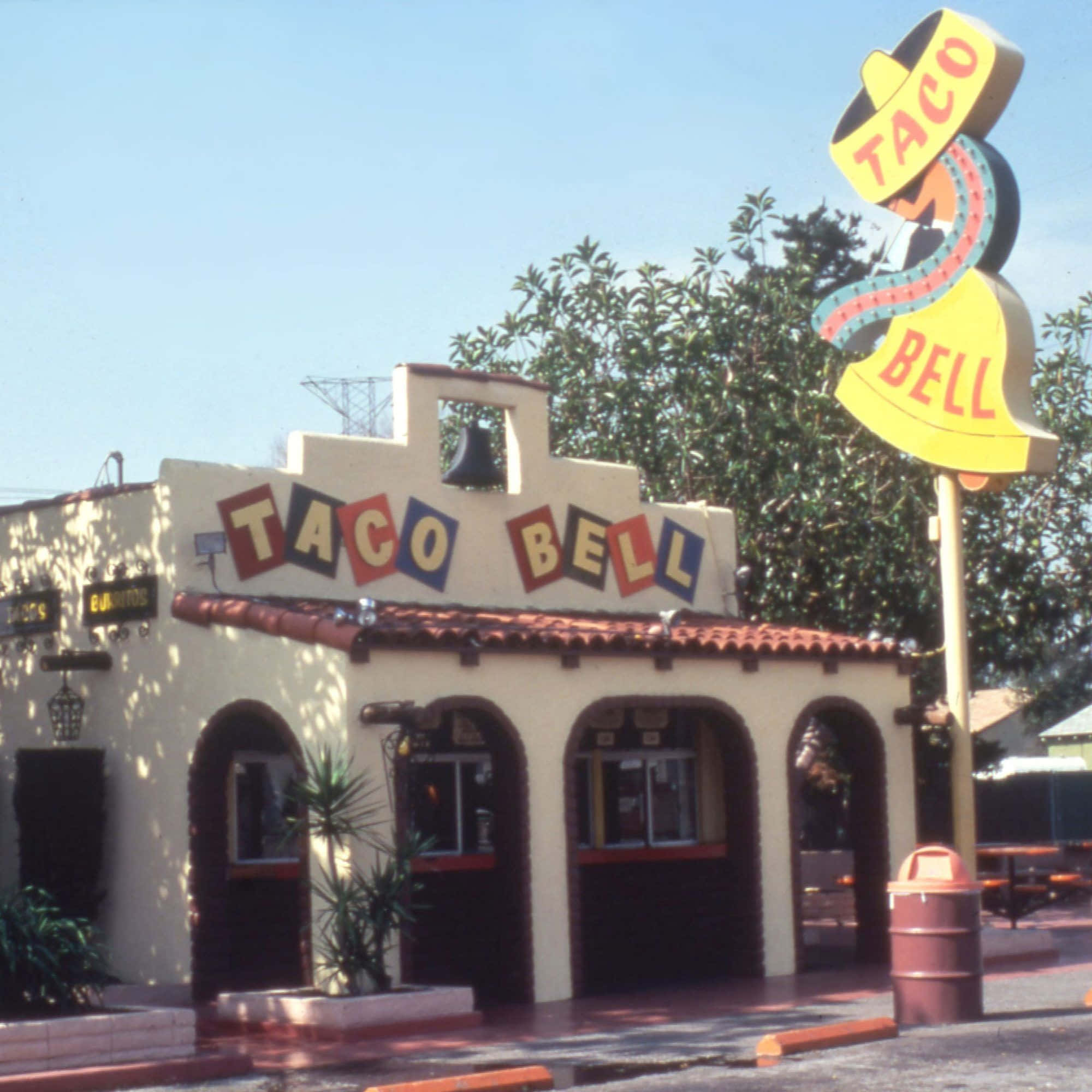塔可鐘 桌布 壁紙 — 一間復古Taco Bell餐廳，在晴朗藍天下有明亮的黃色鐘形招牌和紅色瓦片屋頂。