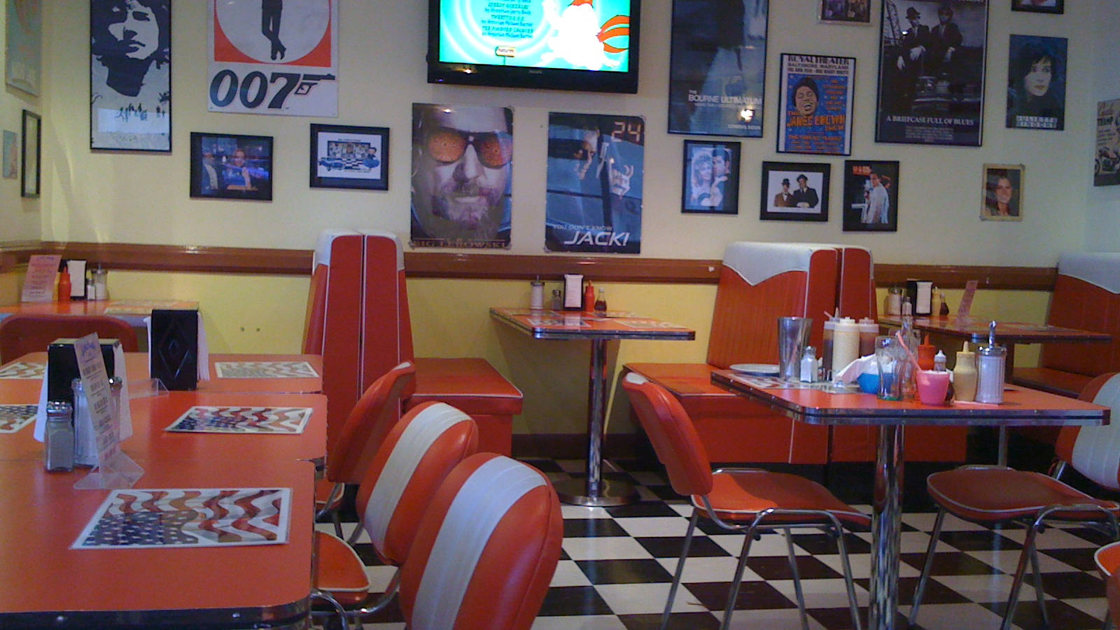 Diner 桌布 壁紙 — 一間復古餐廳，設有橙色卡座、棋盤格地板和黃色牆上的電影海報。