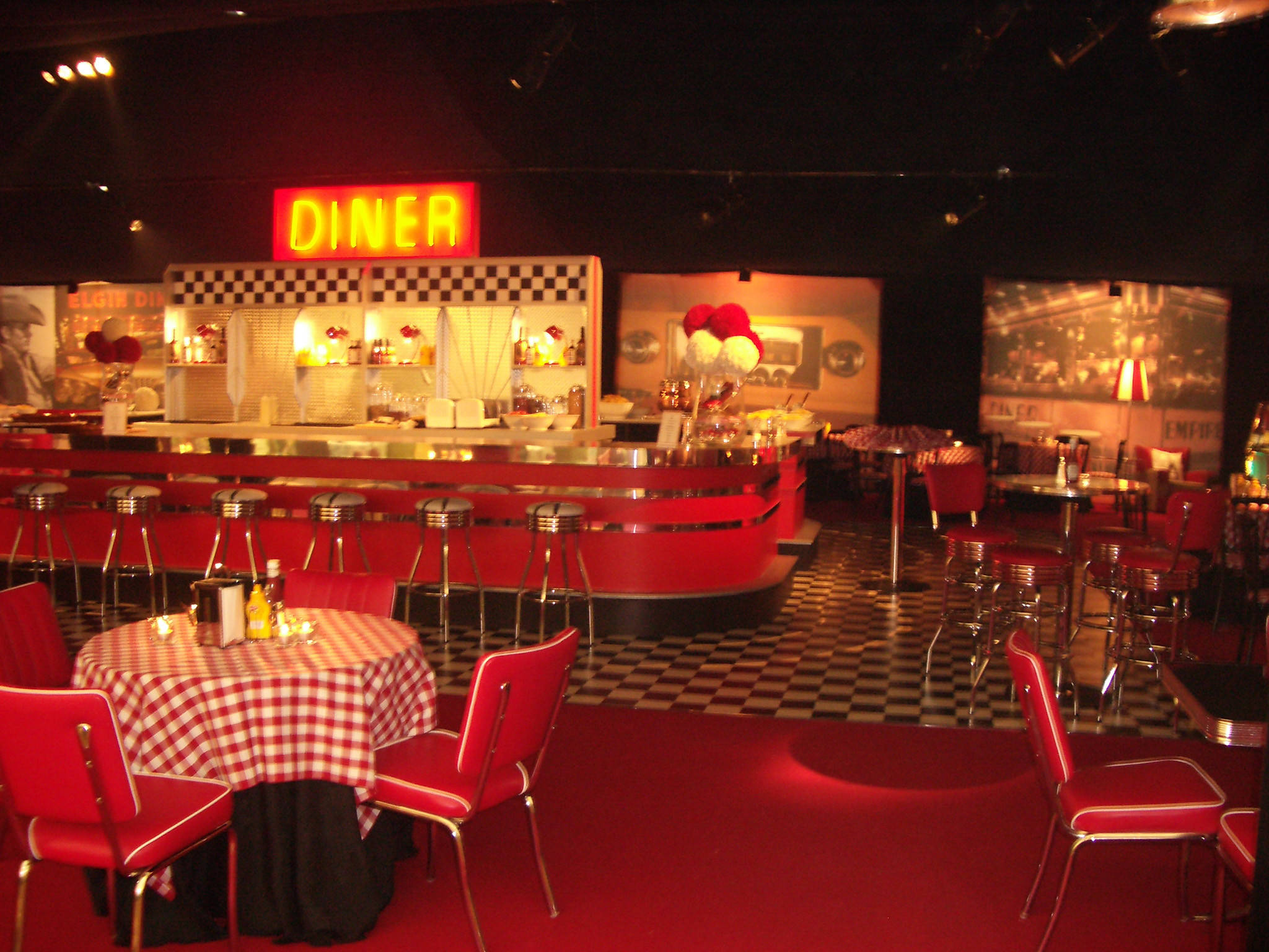 Diner 桌布 壁紙 — 一間復古餐廳，配有紅色乙烯基椅子、棋盤格地板和一個發光的霓虹燈招牌，將溫暖的光線投射在桌子和凳子上。