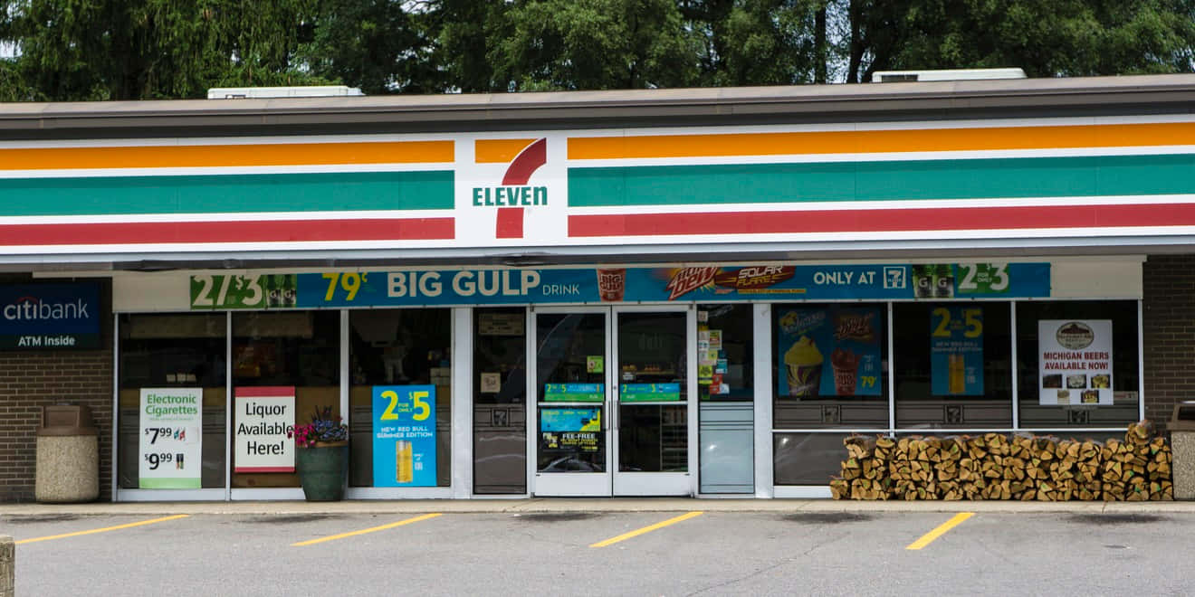 7-ELEVEN 桌布 壁紙 — 一間明亮的7-Eleven便利商店，帶有色彩繽紛的招牌，外面在樹蔭天空下有一堆柴火。