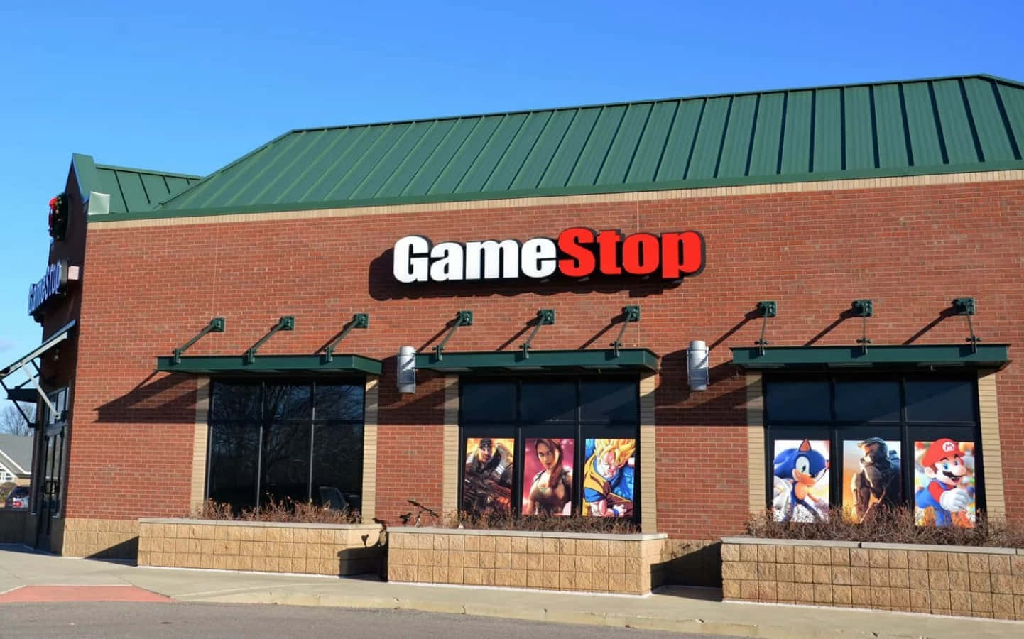 GameStop 桌布 壁紙 — 一間明亮的GameStop商店，磚砌外牆、綠色屋頂，窗戶上貼滿色彩繽紛的電子遊戲角色海報，背景是清澈的藍天。