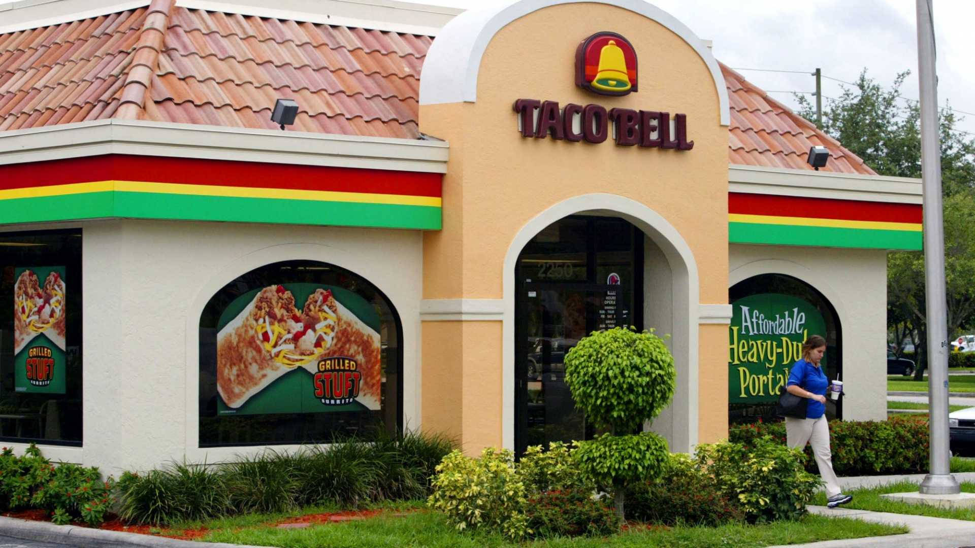 塔可鐘 桌布 壁紙 — 一間明亮的Taco Bell餐廳，帶有紅、黃、綠色飾邊，展示大型食物海報，並有一個人行人在人行道上走過。